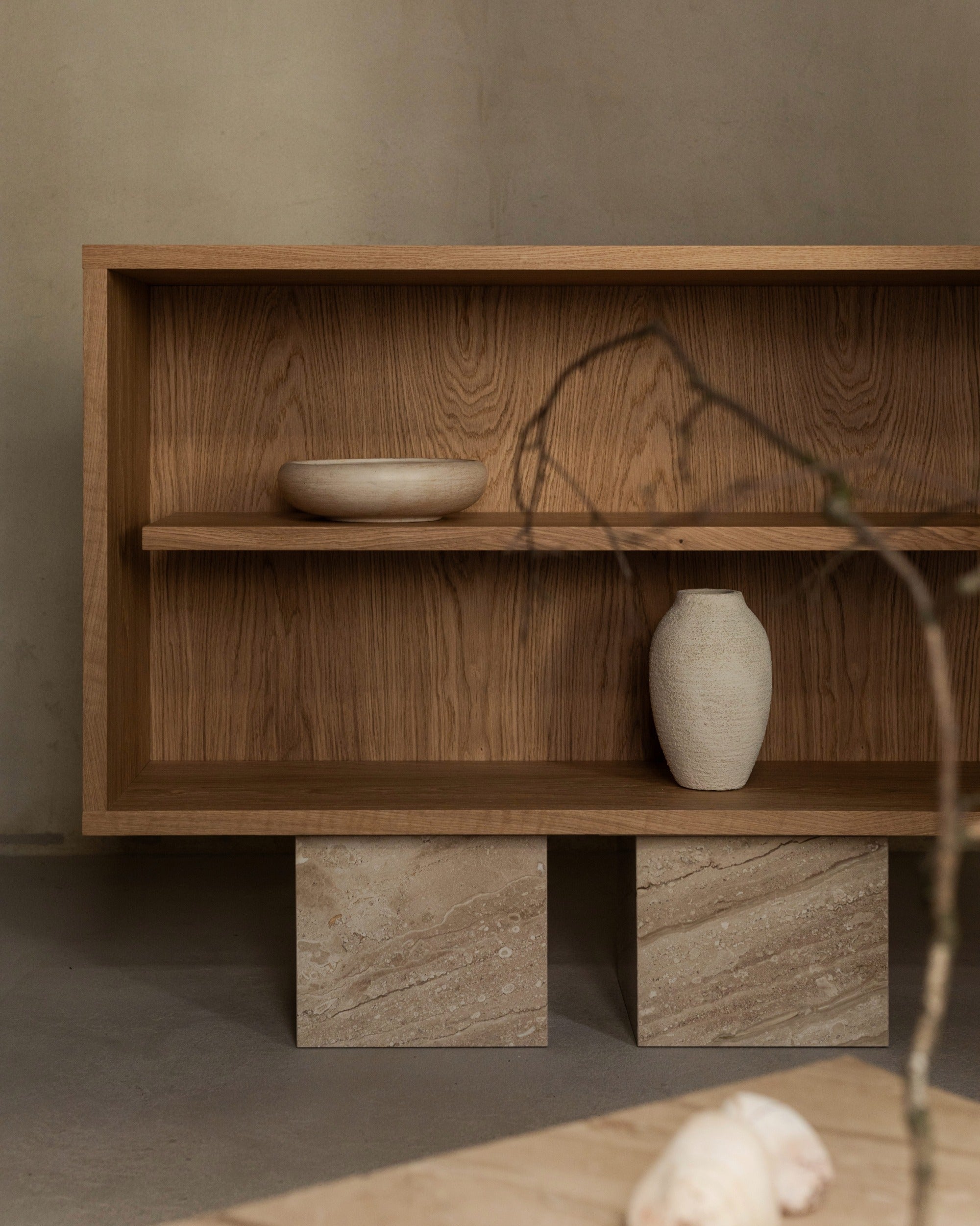 The BLOCK Sideboard by un'common features a minimalist wooden shelf with a round bowl and textured vase, elevated on two stone blocks in a neutral-toned room. A blurred branch in the foreground highlights its elegant design.