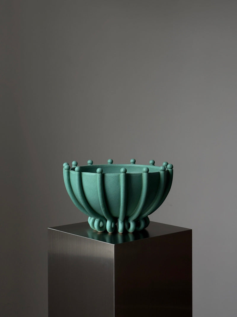 Vessel No. V by Devin Wilde is a green, sculptural bowl featuring raised circular accents and vertical ridges, displayed on a reflective metal pedestal against a plain gray background.