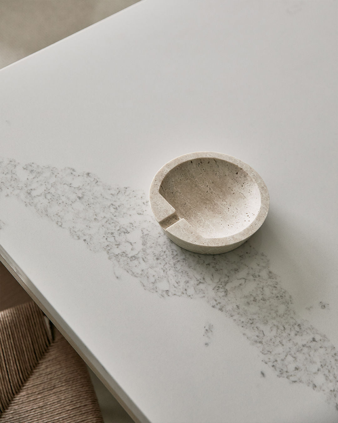 The JCL Ashtray in Travertine Stone, round and speckled with a notch, rests on a subtly veined light gray marble surface. A striped beige chair appears in the lower left corner.