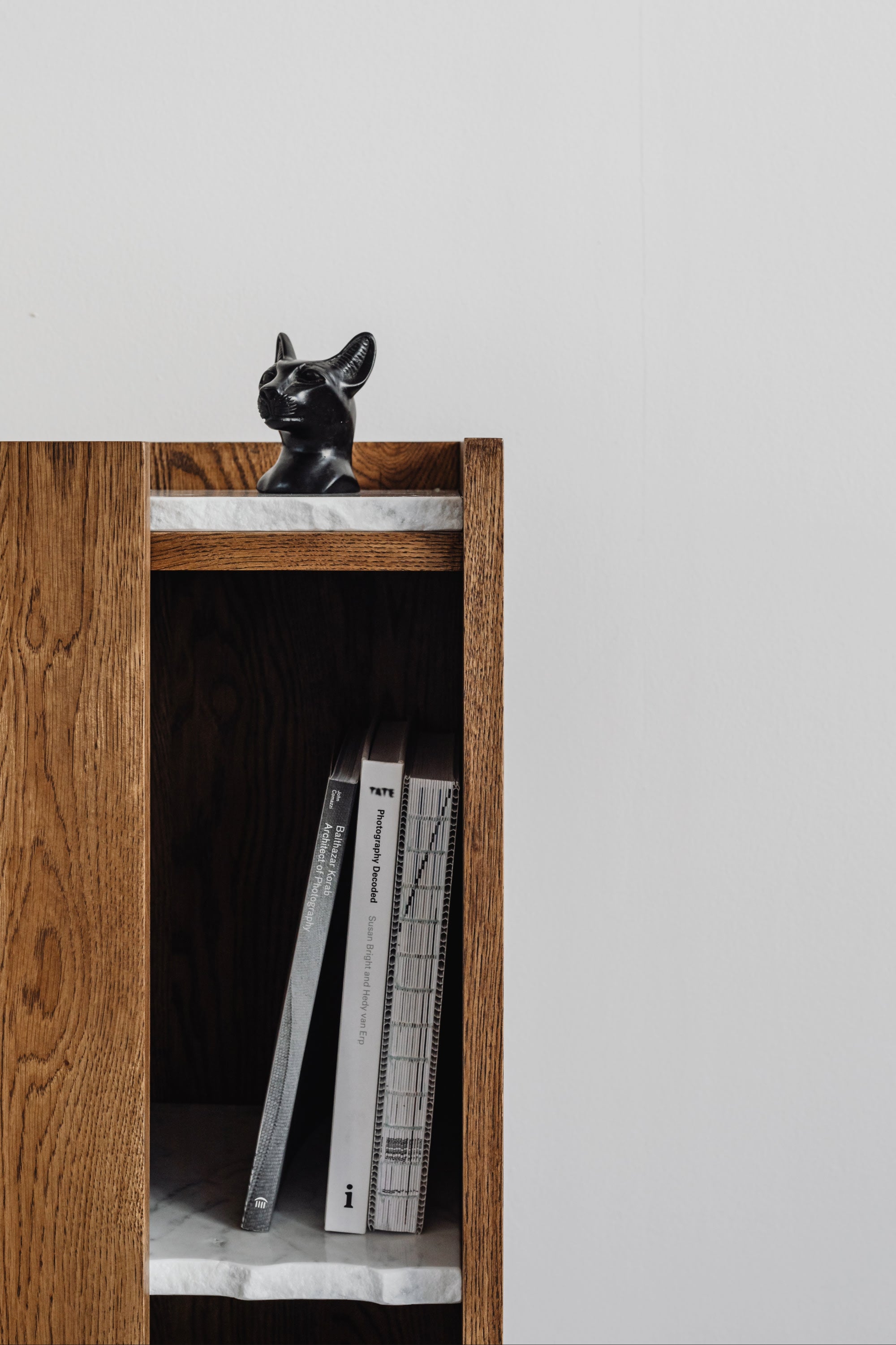 The PAVO Cabinet by un'common features a white marble shelf displaying three vertically stacked books and a small black cat figurine, all set against a plain white wall.