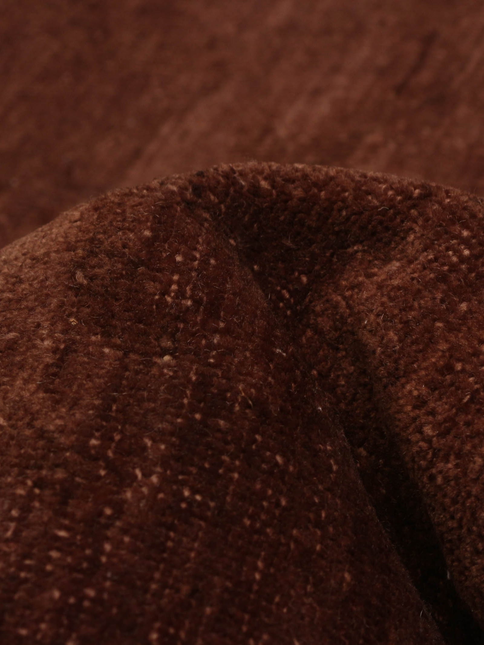 Close-up of the soft, textured Overdyed Rug in Russet by catalog:three (178 x 277 cm), with a slightly folded appearance. Its surface features a subtle weave pattern, enhancing its rich, earthy color.