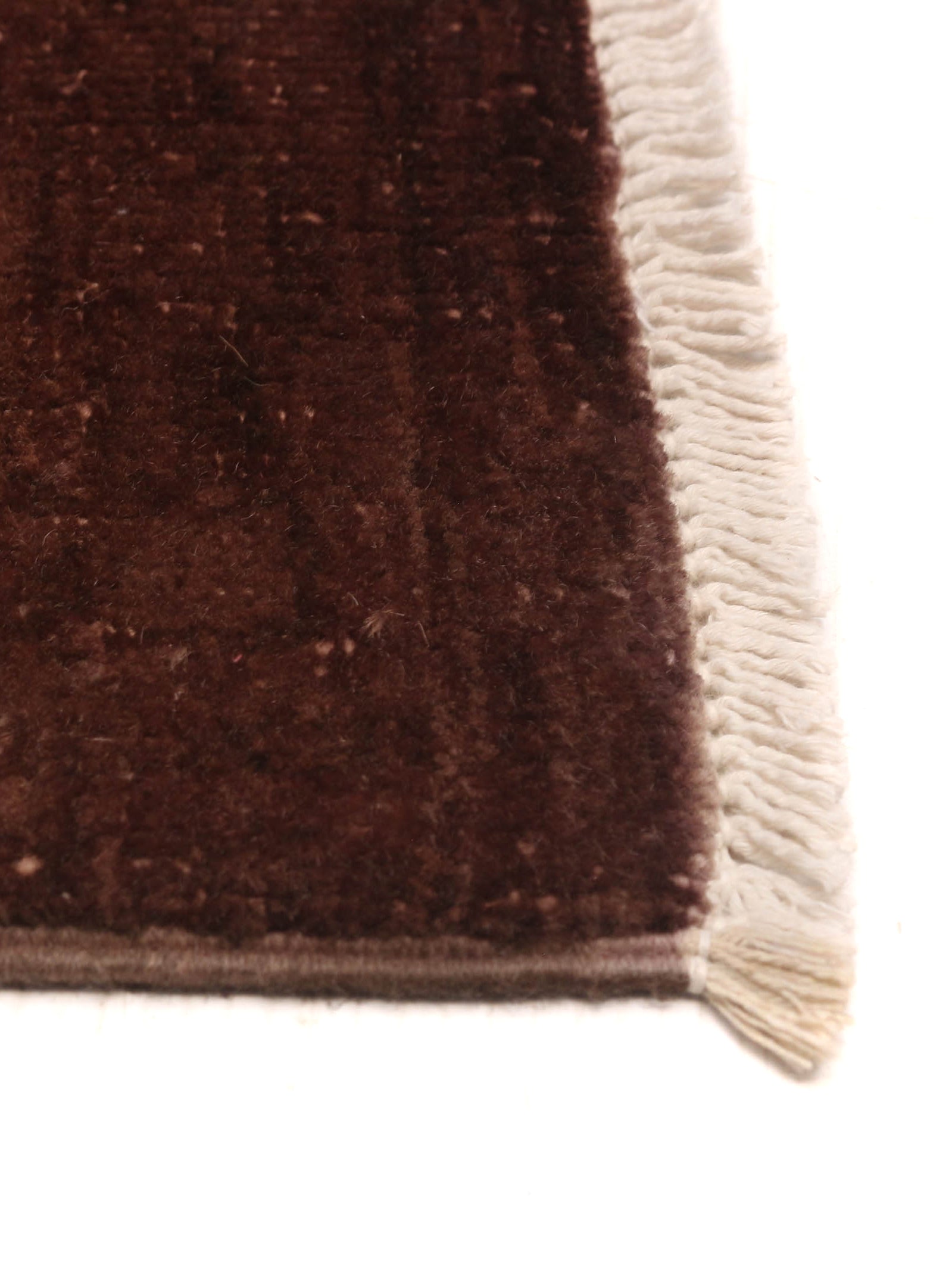 Close-up of the corner of the Overdyed Rug in Russet by catalog:three, featuring a dark brown wool texture with a cream border and fringed edge on a white background.