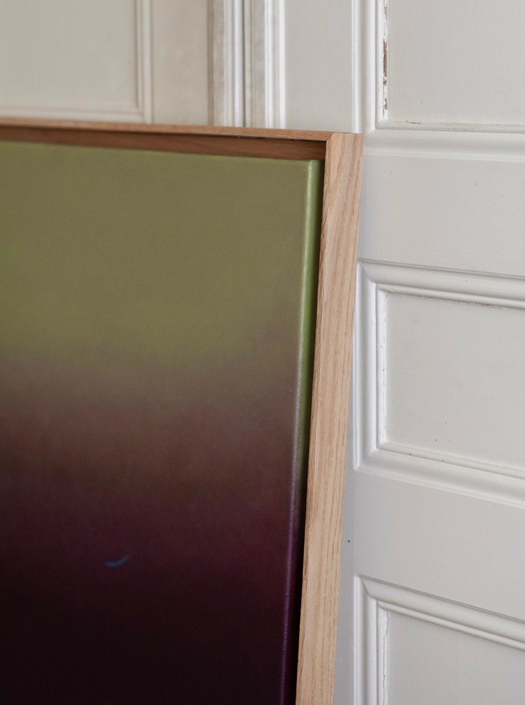 A close-up image showing Something(s) in the air, 2024 by Rebecca von Matérn, featuring a gradient-colored artwork leaning against a white, paneled wall. The piece shifts from green at the top to dark maroon at the bottom, with a hard waxed oak frame partially in view.