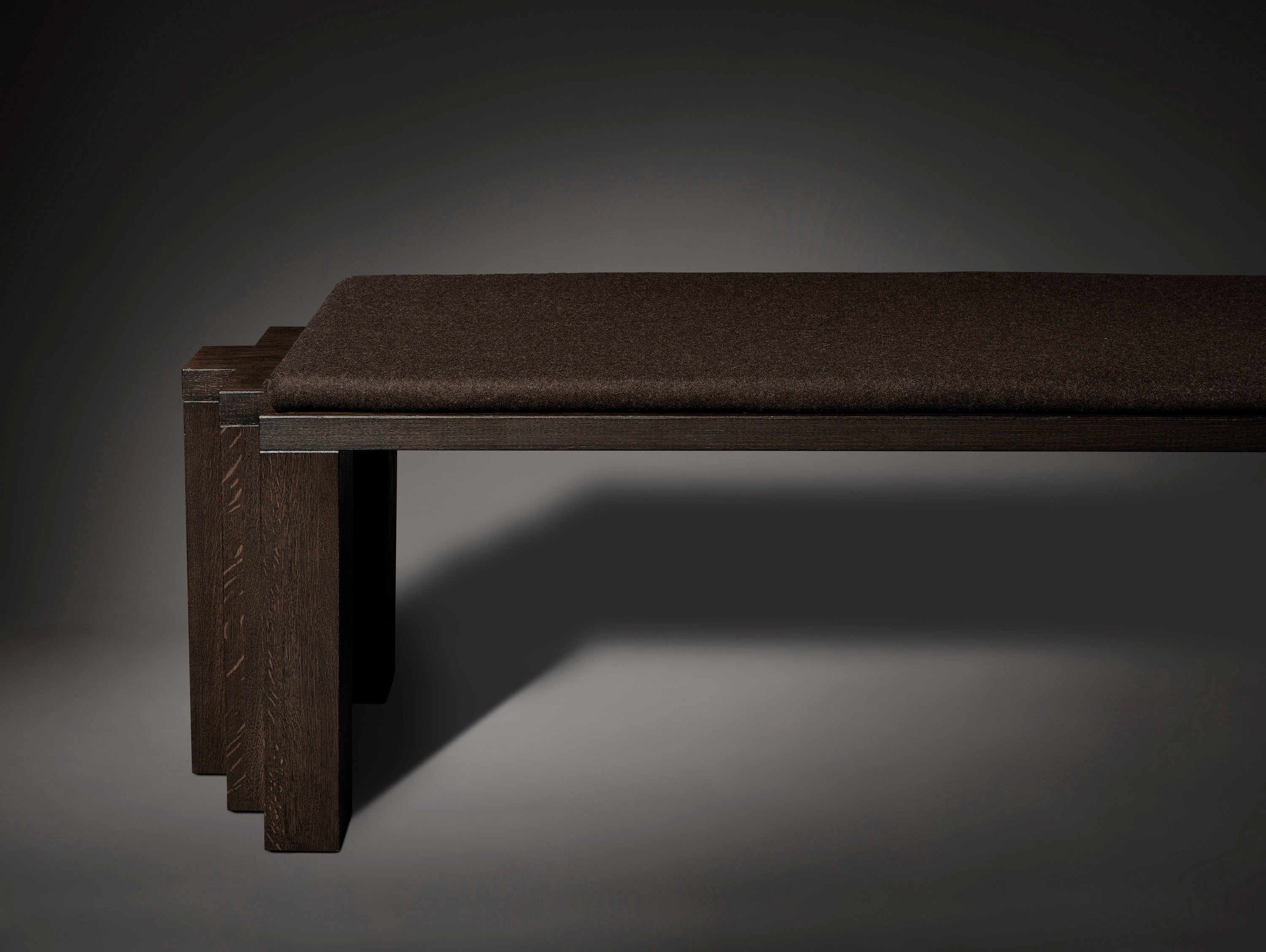 The Mokko Cadence Bench, a minimalist piece made from solid European oak with thick rectangular legs and a flat cushioned seat, is shown partially against a plain dark background. Only the legs and seat of this customizable bench are visible.