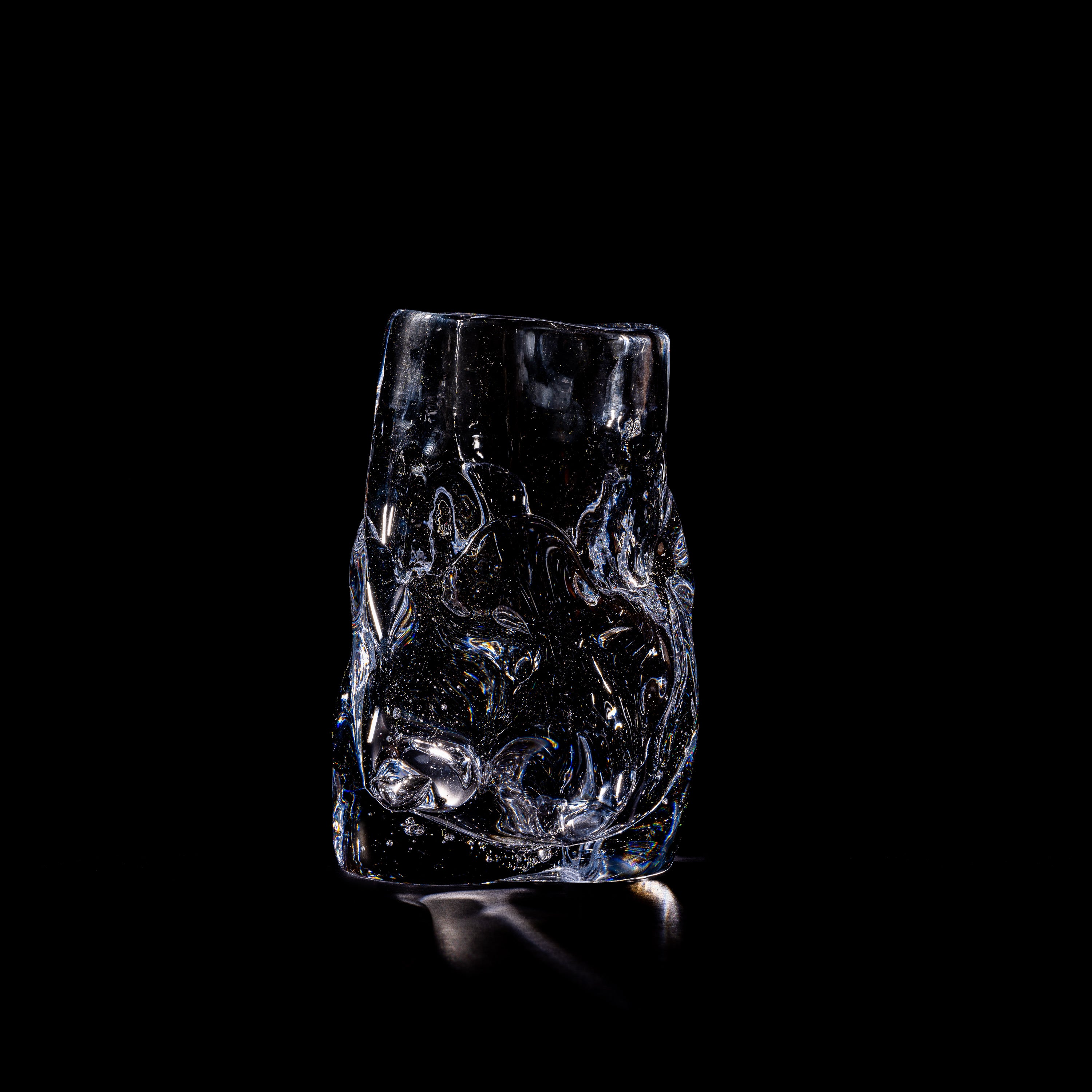 Szkło Studio's Organically Shaped Hand-blown Vase No. 2 is illuminated against a black background, highlighting its unique organic form and textured surface reminiscent of handcrafted glass art. Bubbles within add depth and intrigue to the piece.
