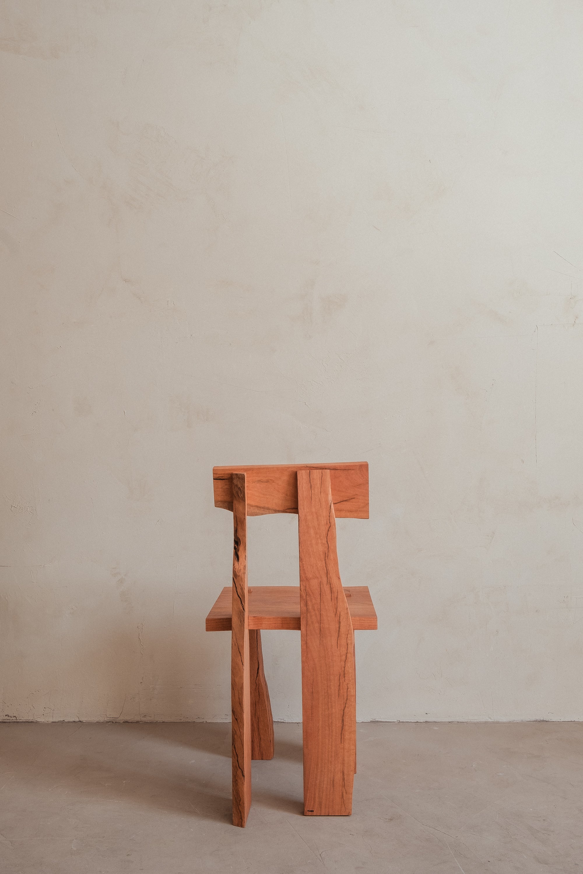 The Askew Chair by Ther features a geometric, angular wooden design. Positioned on a bare concrete floor against a light, textured wall, it highlights modern minimalist elegance.