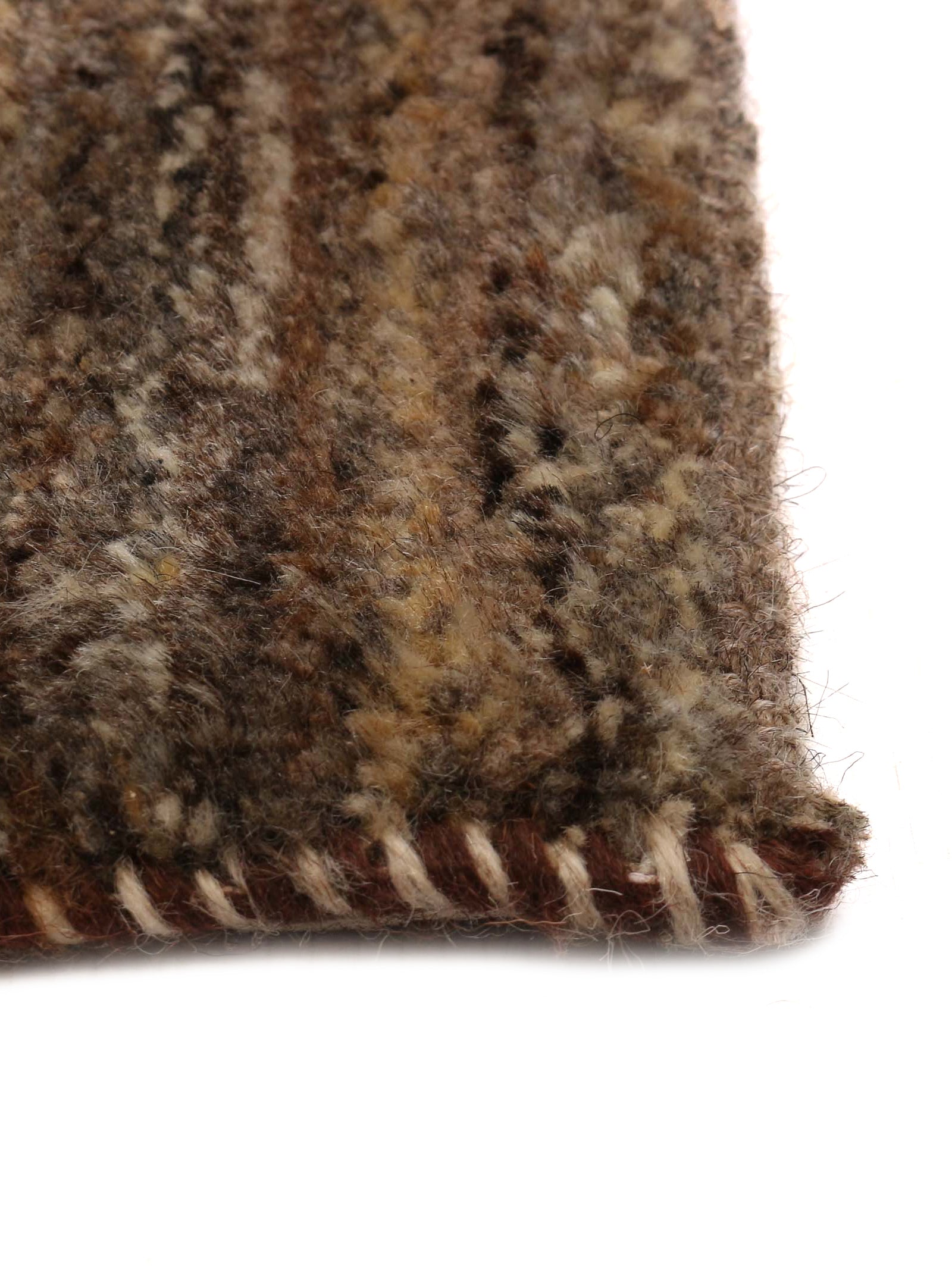 Close-up of a corner of the Gabbeh Rug in Sable by catalog:three, measuring 174 x 232 cm. This unique piece features a thick, plush woolly texture with multicolored shades of brown, beige, and gray. The edge is neatly finished with visible contrasting stitching.