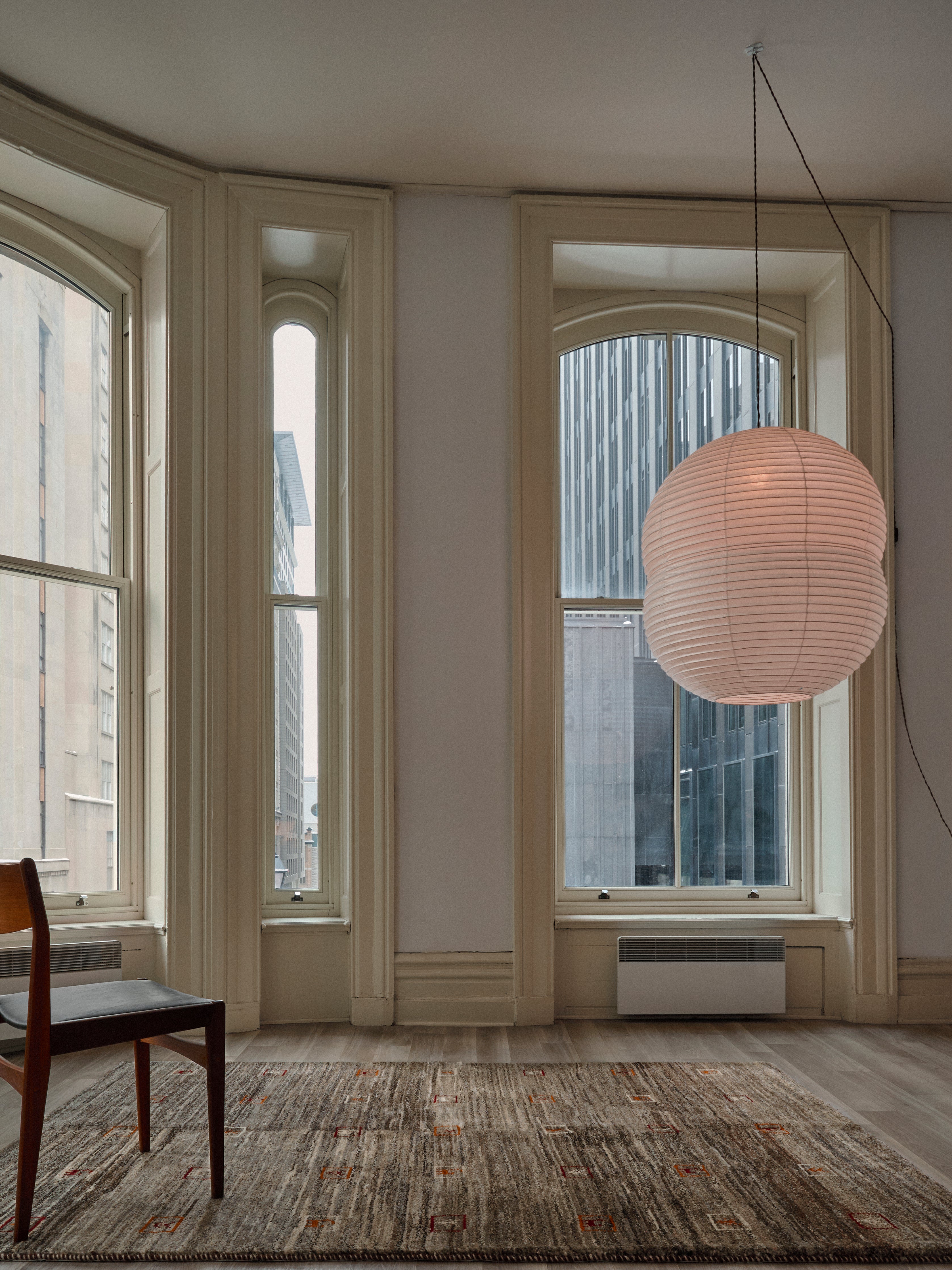 The room features large arched windows with city skyscrapers, a wooden chair, and a round paper lantern. The light-colored walls accentuate the spaciousness, while the floor is adorned with a plush thick pile Gabbeh Rug in Sable (174 x 232 cm) from catalog:three.