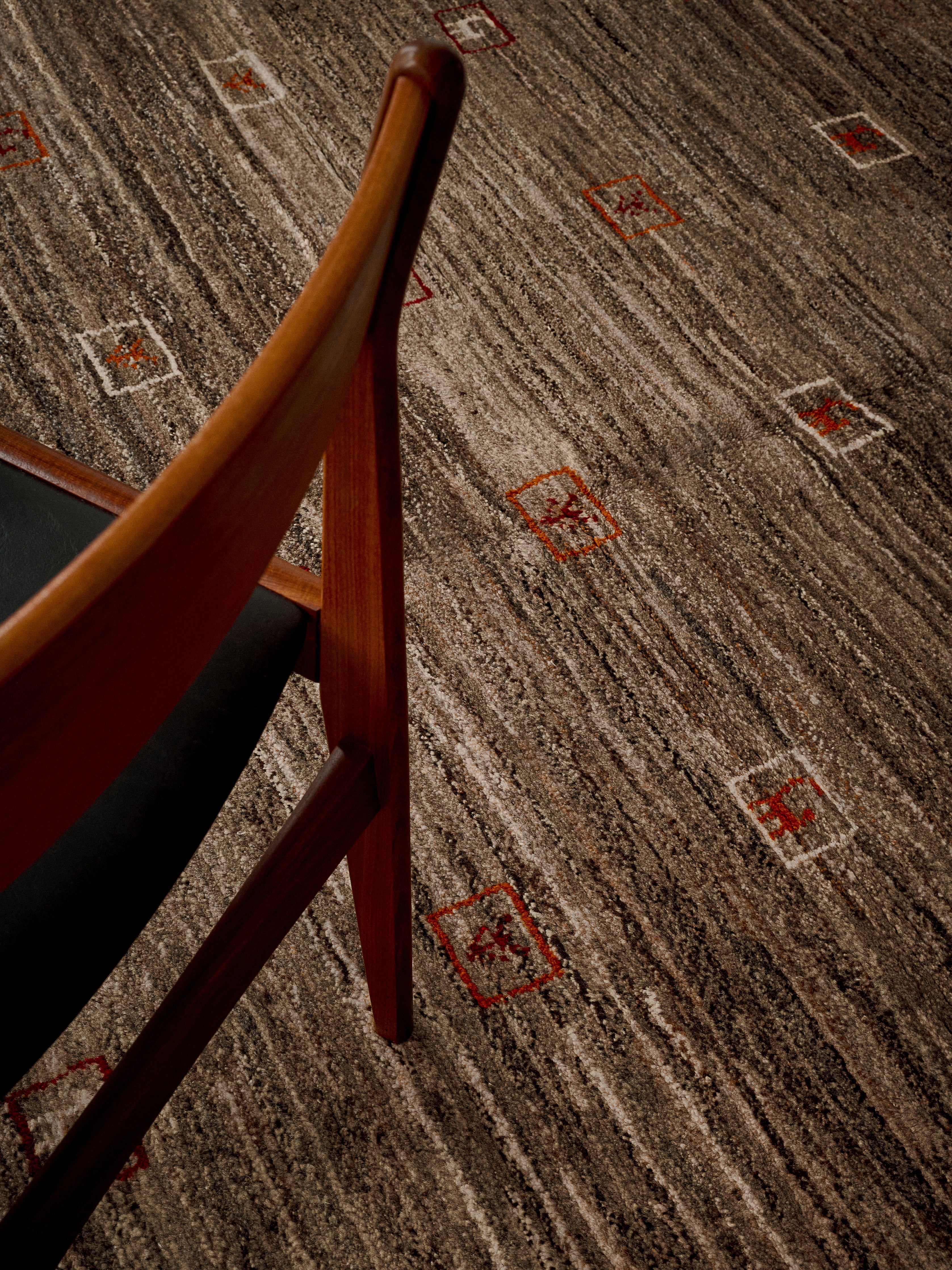 A wooden chair with a black seat rests on the Gabbeh Rug in Sable, intricately woven in India by catalog:three, featuring small red and white squares.