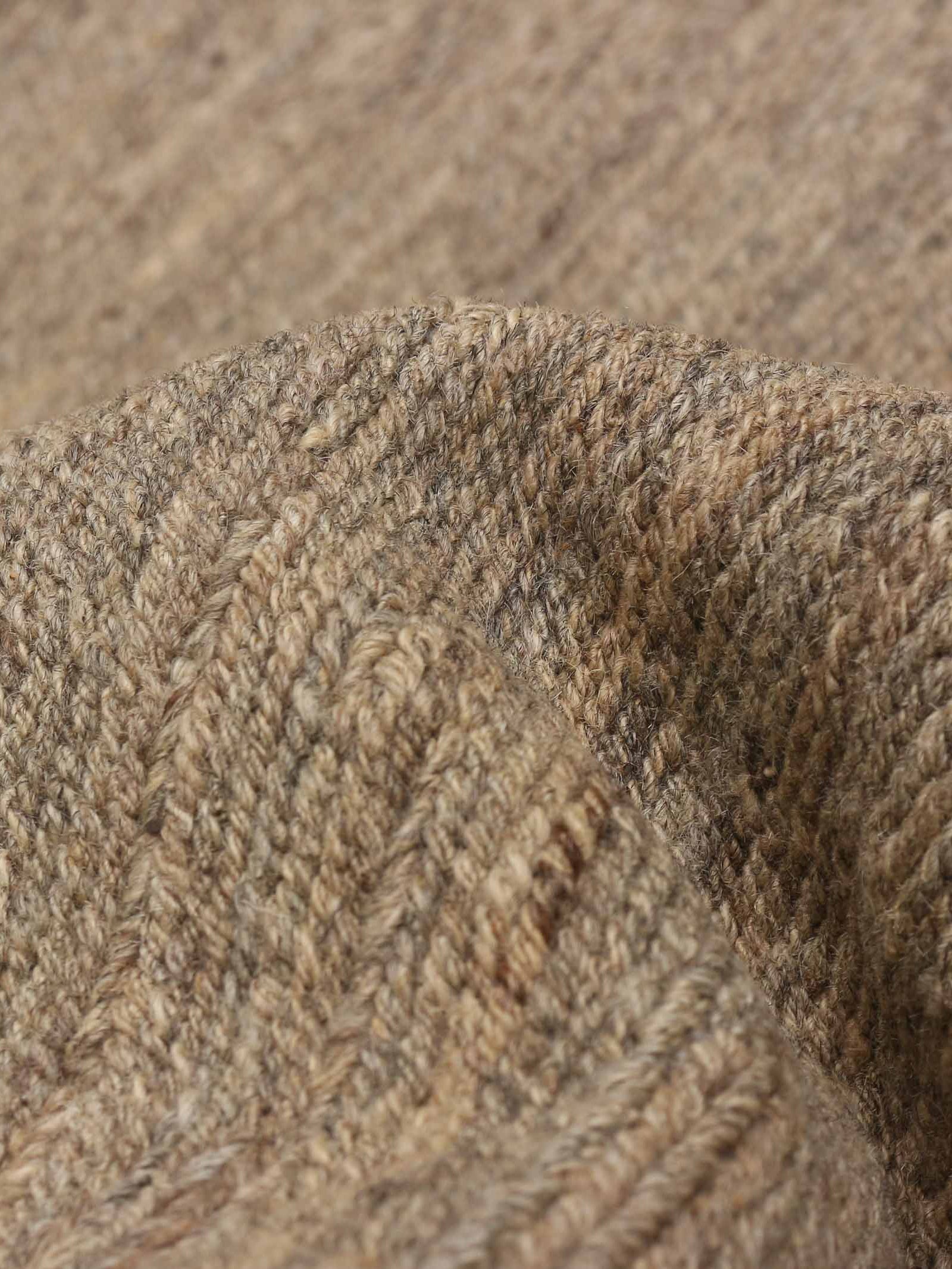 A close-up of the Flatweave Rug in Sandalwood by catalog:three reveals a soft, textured surface with a subtle pattern like a hand-woven kilim. The intertwining brown and beige fibers create a diagonal weave reminiscent of Pakistani craftsmanship. Dimensions: 238 x 300 cm.