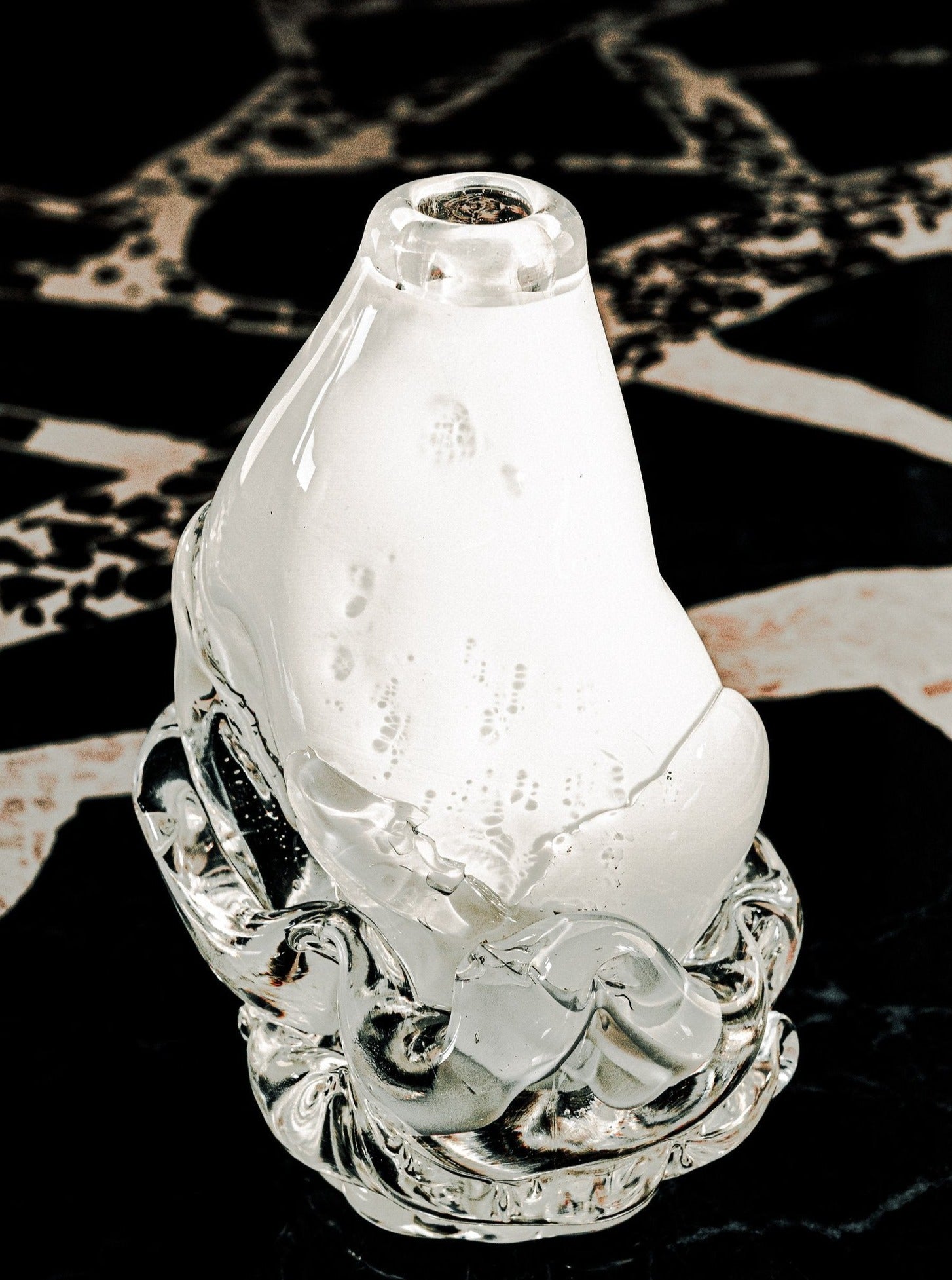 A close-up of the Szkło Studio Opal Vase - Small, a handcrafted clear glass piece with a rounded top, swirls at the base, sculptural bubbles, and internal textures, showcased on a dark patterned surface.