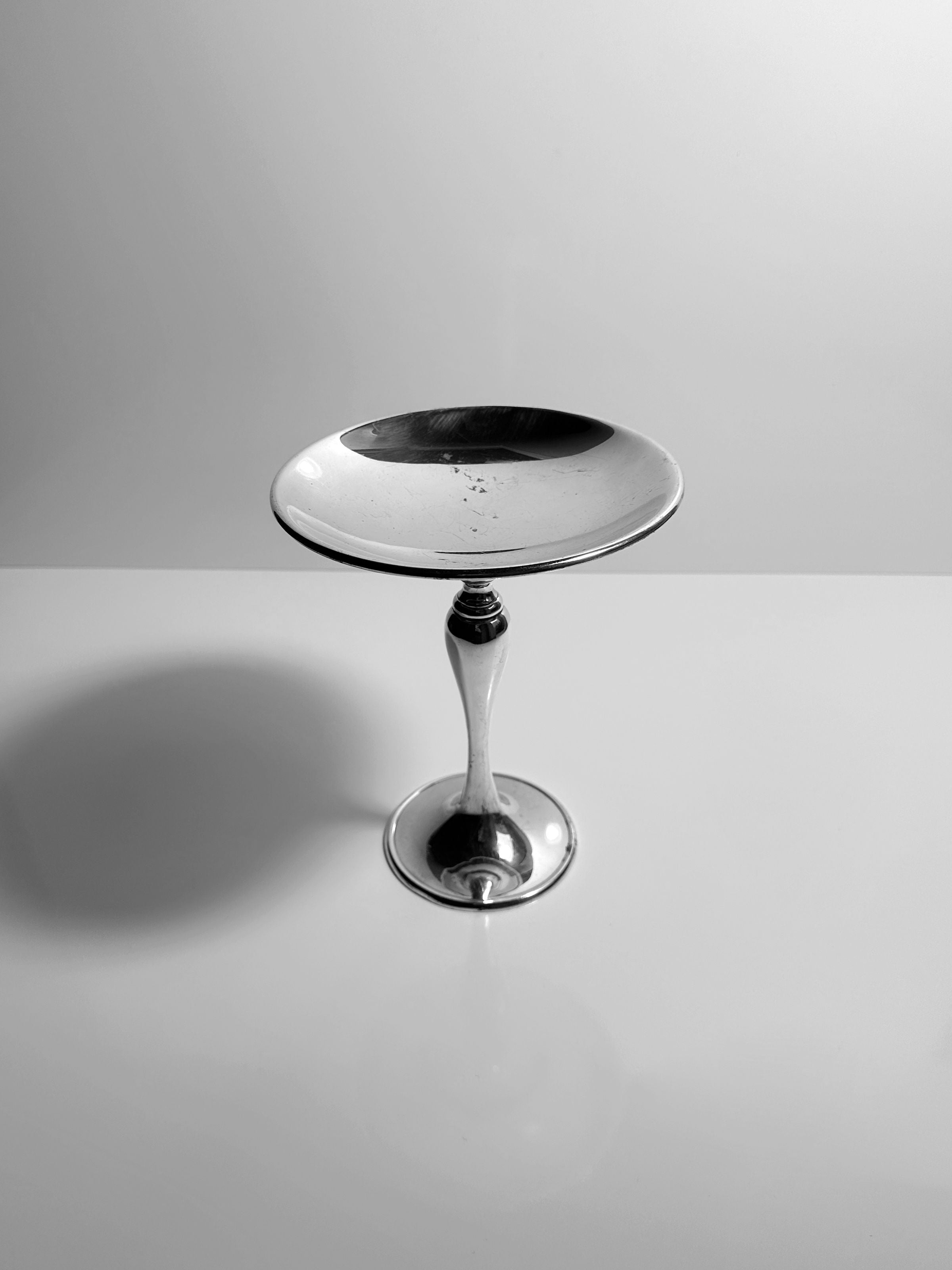 The Dodo Vintage Heavy Silver-Plated Foot Bowl, with its wide, shallow design and gleaming finish, sits on a reflective white surface and casts a soft shadow leftward against a plain light grey background.