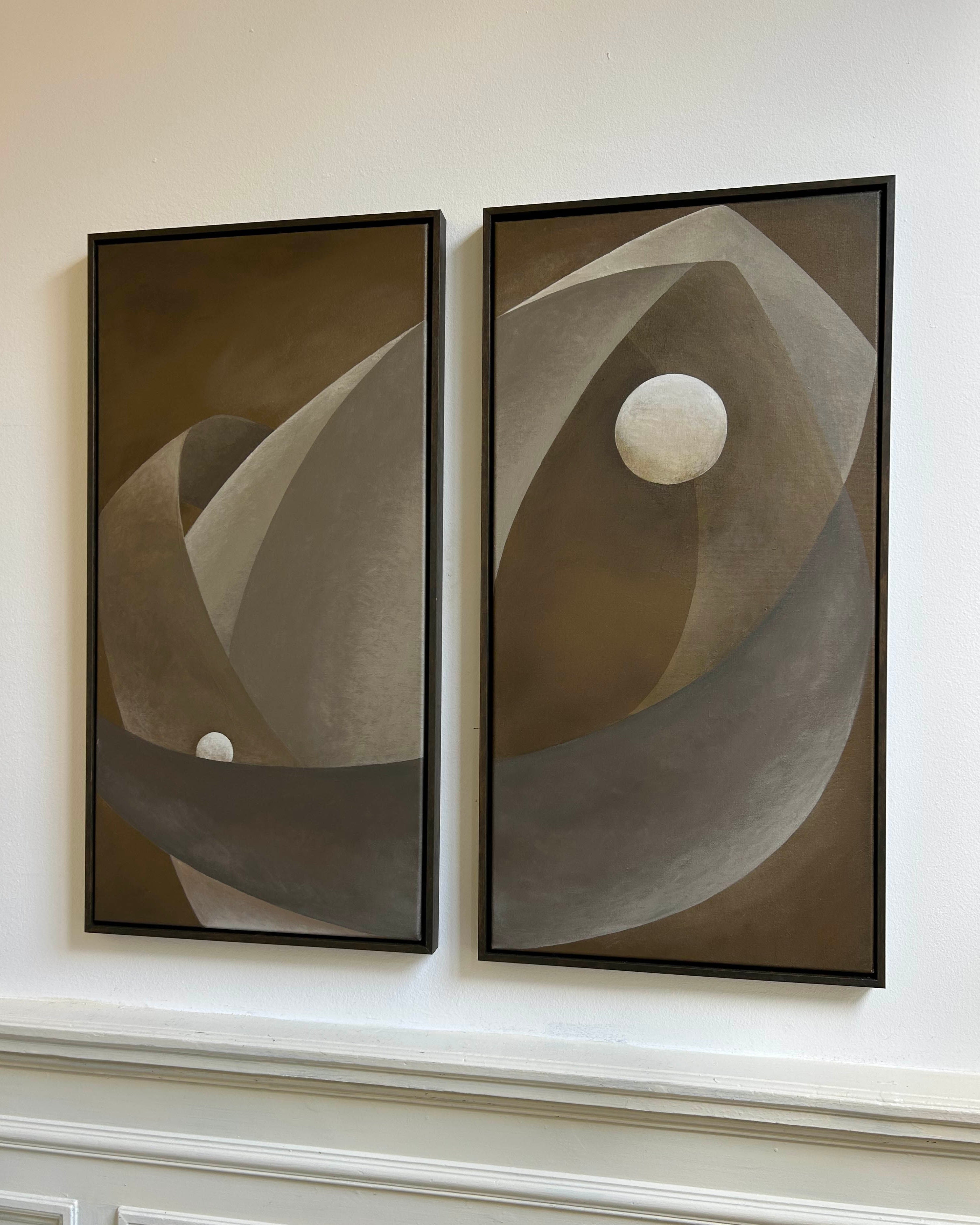 The 'You only live twice' paintings by Iris Bavelaar, featuring abstract curved shapes and white circles in brown, beige, and gray on canvas and linen, are displayed side by side above decorative molding on a white wall.