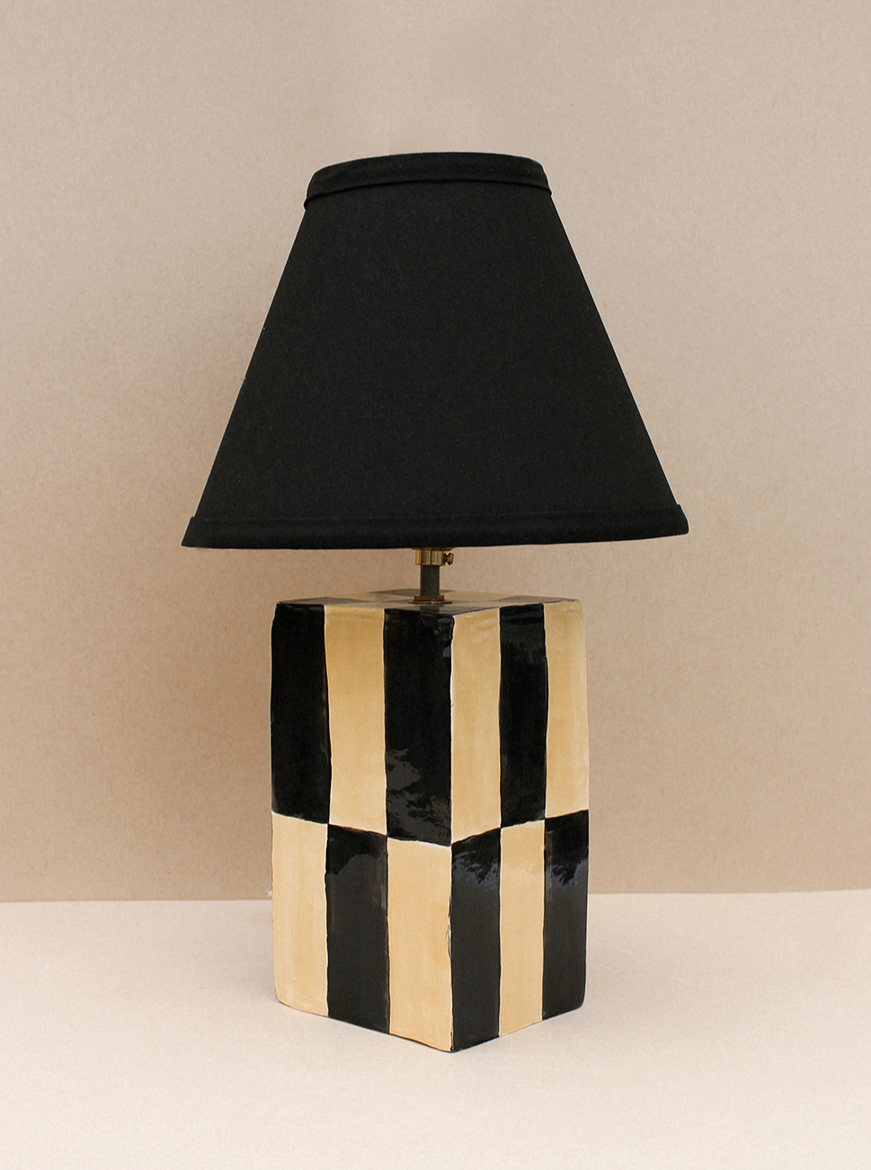 A Socorro Lamp with a black lampshade, mounted on a square base decorated with black and beige vertical stripes against a plain beige background. (Brand Name: Casa Veronica)