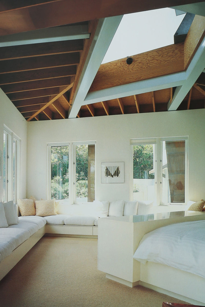 A bright, modern bedroom with a large corner sofa, neutral tones, exposed beams, and a skylight. Vintage books and the Villa Architecture Book Series - Set of 5 add character, while multiple windows bring in natural light.