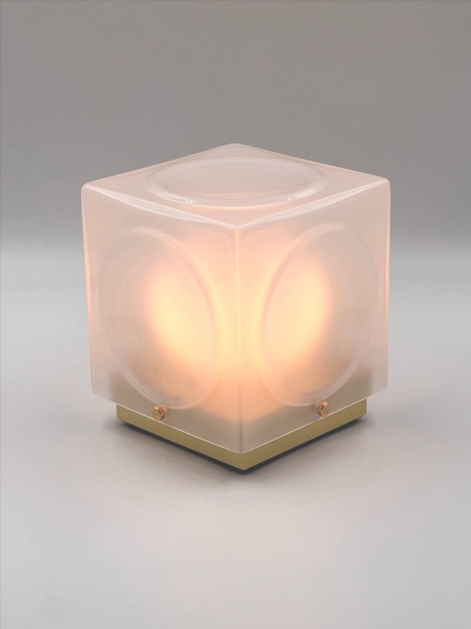 The Chant Portable Table Lamp by Lee Broom features frosted translucent sides and smooth, rounded corners on a square base, softly glowing with warm light against a neutral background.