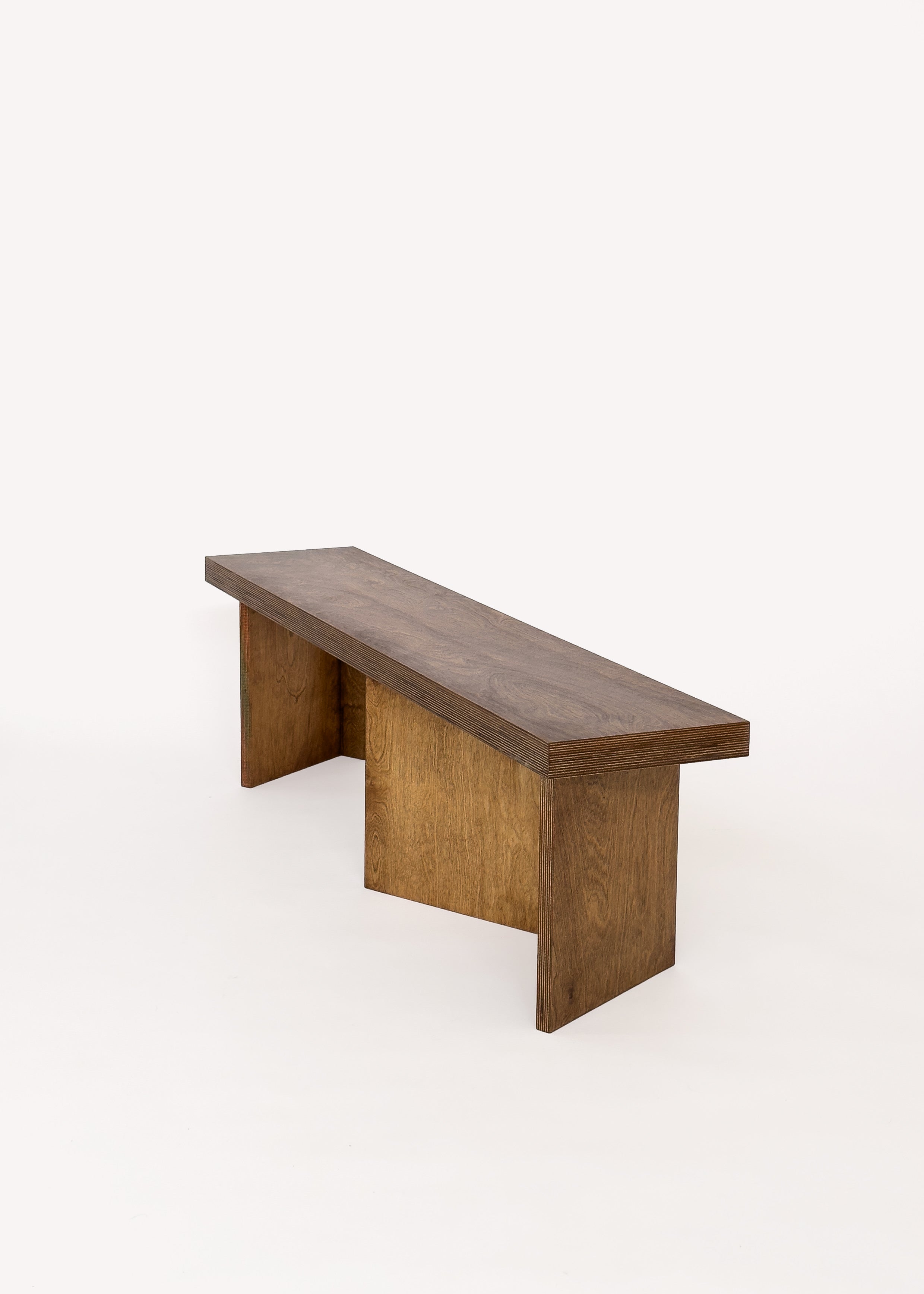 The Free plan Bench by Goons from GOONS features a minimalist rectangular wooden seat and two wide, flat legs in a medium brown finish—modern, architectural furniture set against a plain white background.