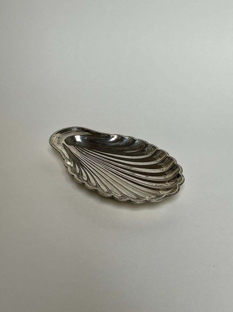 The Manushi Silver Shell Dish, shaped like a scallop with detailed ridges and a polished, reflective surface, rests on a plain light gray background.