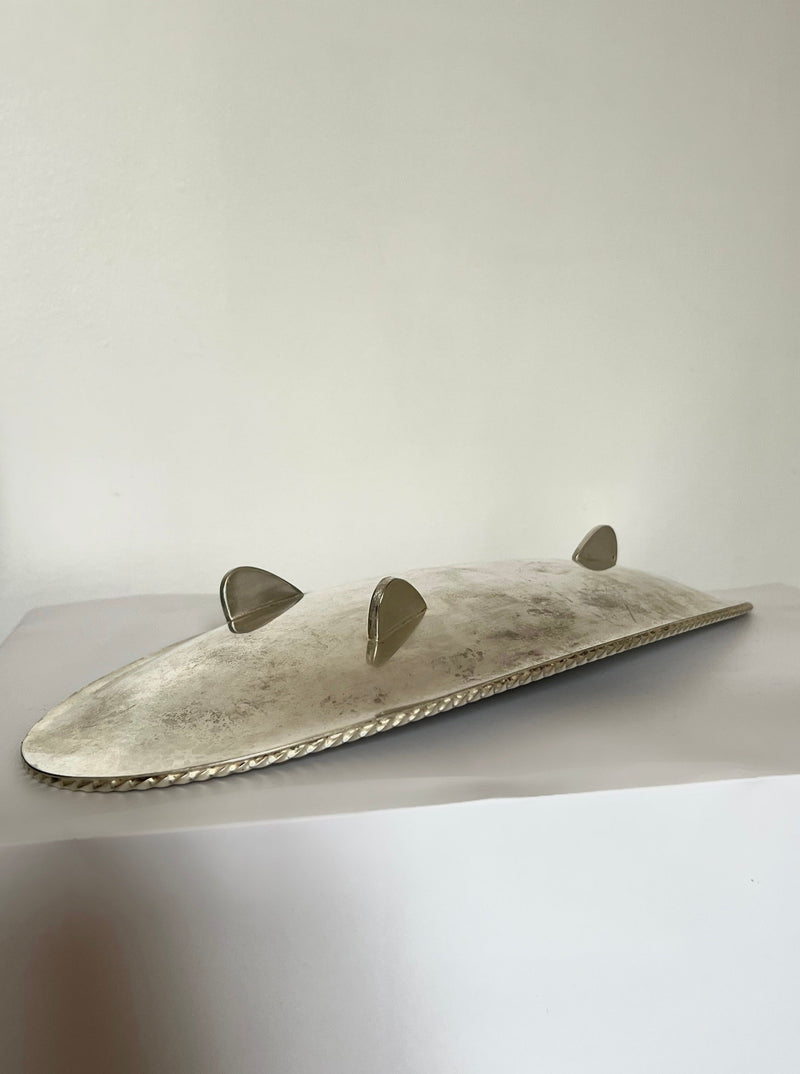 The Manushi Long Silver Serving Dish features an oval shape, textured edge, and three curved handles, shown resting on a white surface against a plain light background.