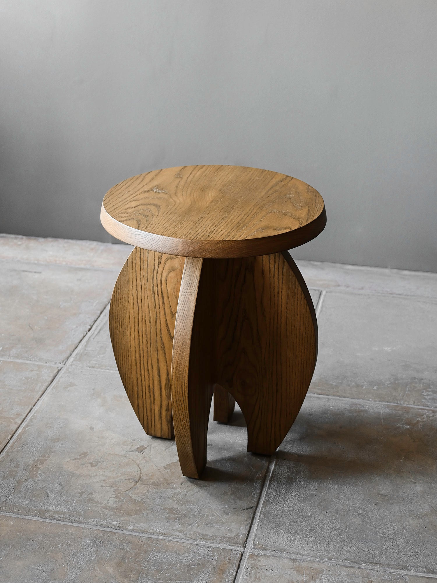 The 'Fusée' Stained Oak Side Table by Studio OSKLO features a small, round design with three curved legs and a natural wood finish that highlights the grain, set on a gray tiled floor against a plain gray wall.
