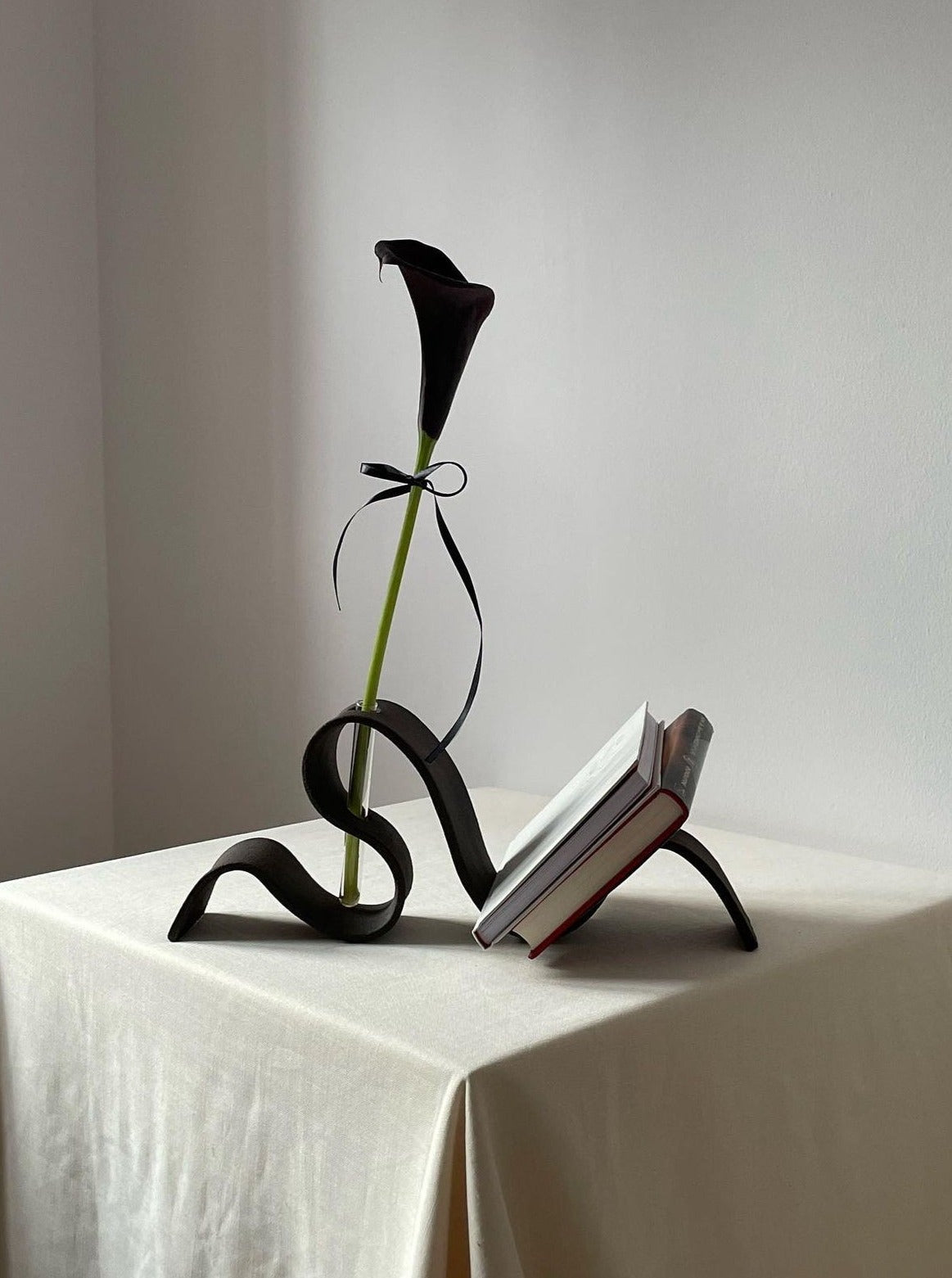 A minimalist table setting shows a single dark calla lily in the Ribbon Vase in Black by LESS, which doubles as a book holder supporting an open book on a light tablecloth against a plain white wall.
