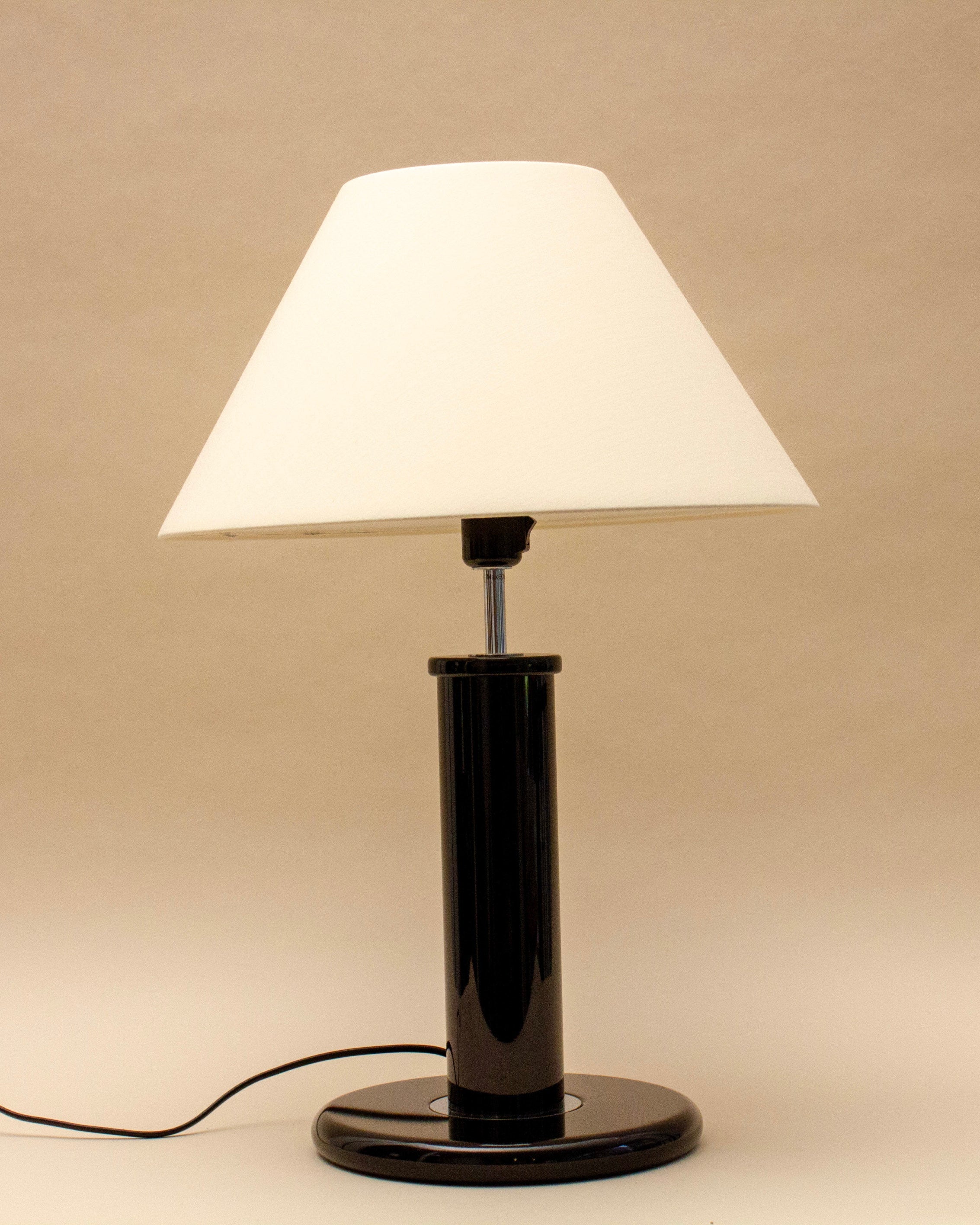 The SÒLER MADRID TABLE LAMP features a white conical shade and glossy black cylindrical base, set on a beige surface with a neutral backdrop. Its visible power cord reflects a blend of classic and modern design elements.