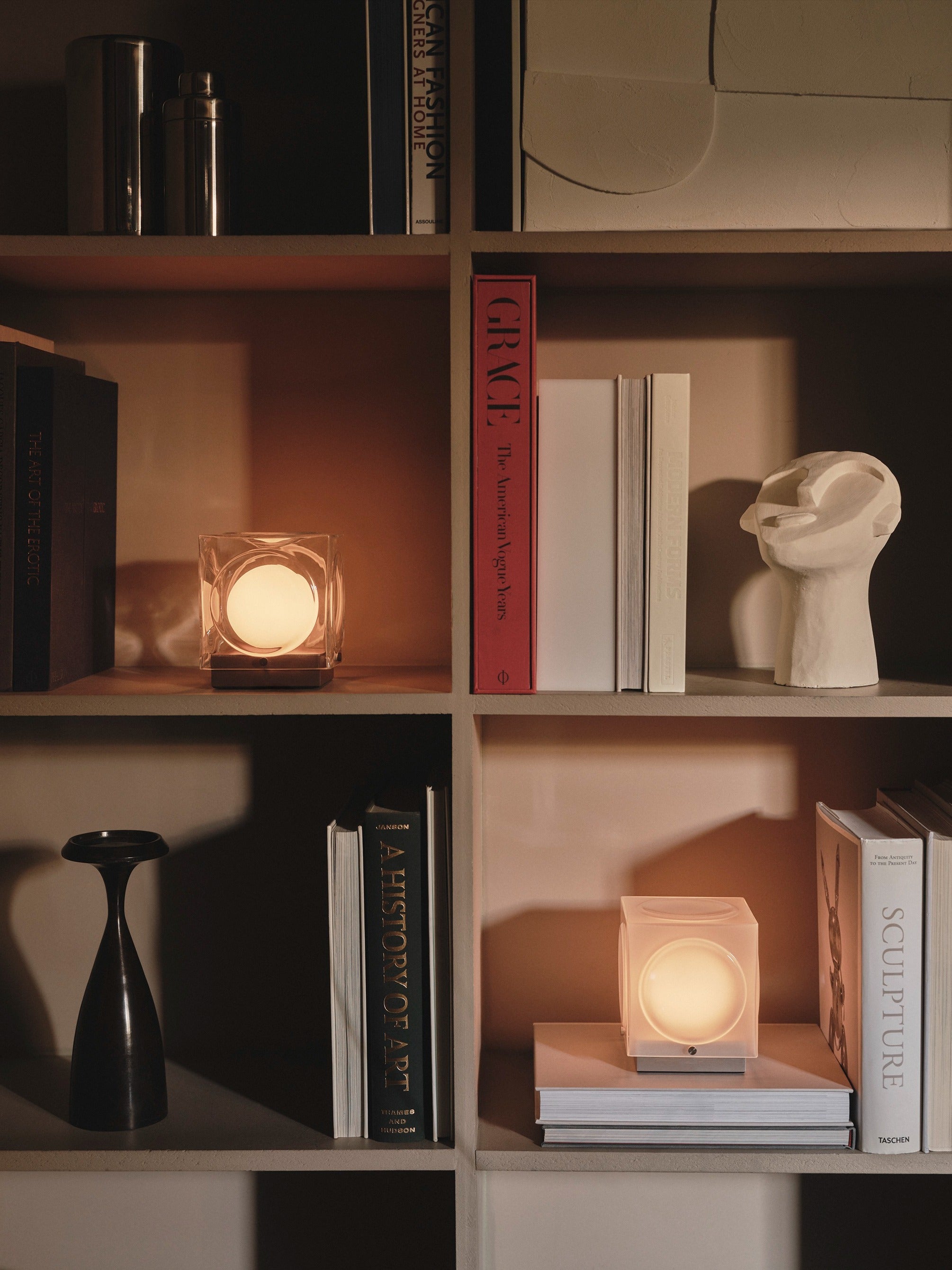A modern bookshelf displays books, the Lee Broom Chant Portable Table Lamp, a warm wireless lamp, a black abstract vase, and a white abstract head sculpture all arranged neatly on the shelves.