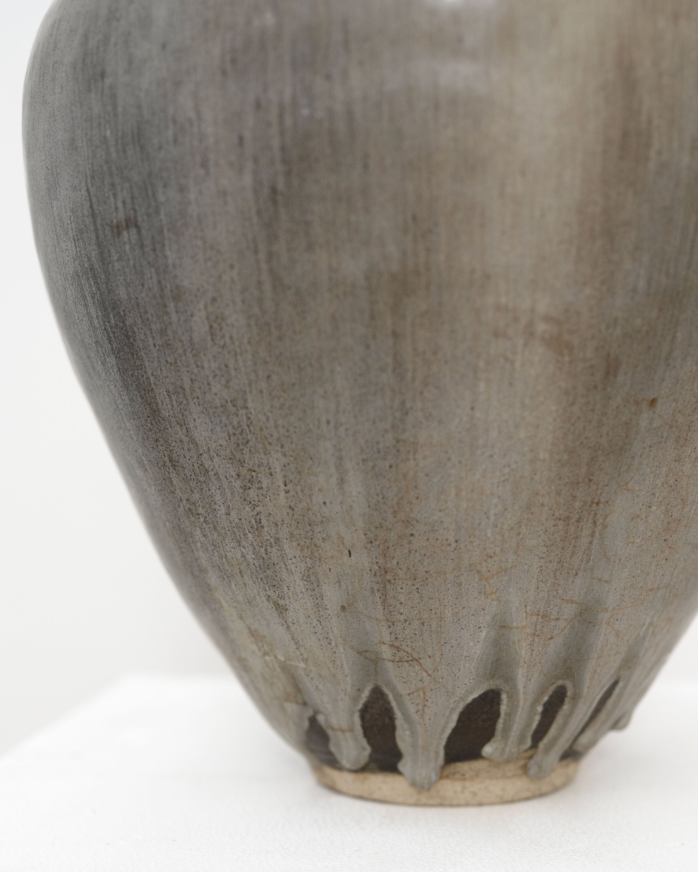 Close-up of the lower half of the Tom Bogle Hand Built Studio Pottery Vase, featuring a smooth grayish surface with subtle brown streaks and white glaze drips down toward its base, resting on a white surface.