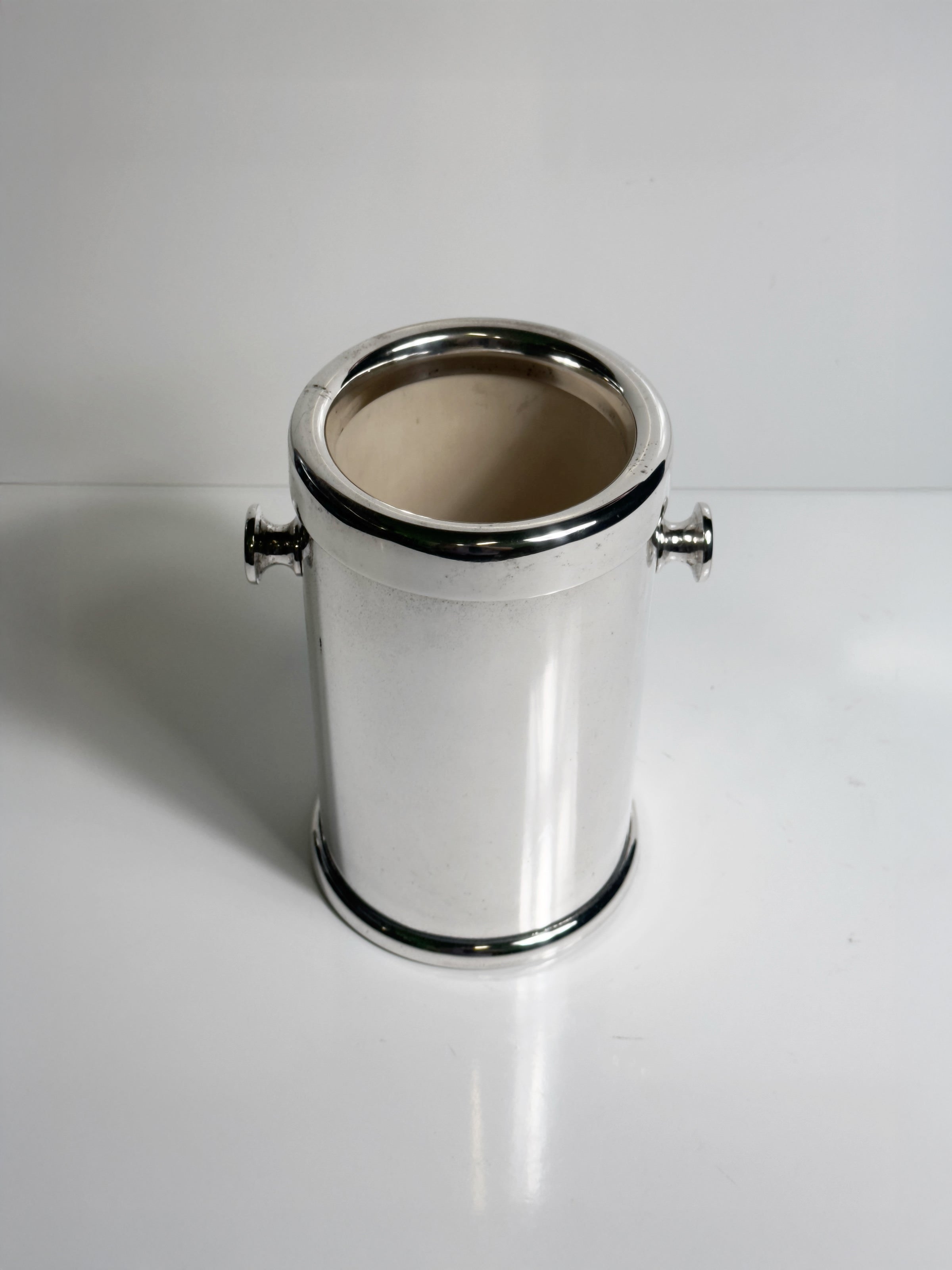The Dodo Vintage Silver Plated Champagne Cooler - 1980s features a shiny, cylindrical design with two small side handles. Shown empty, it sits on a white surface against a plain white background.