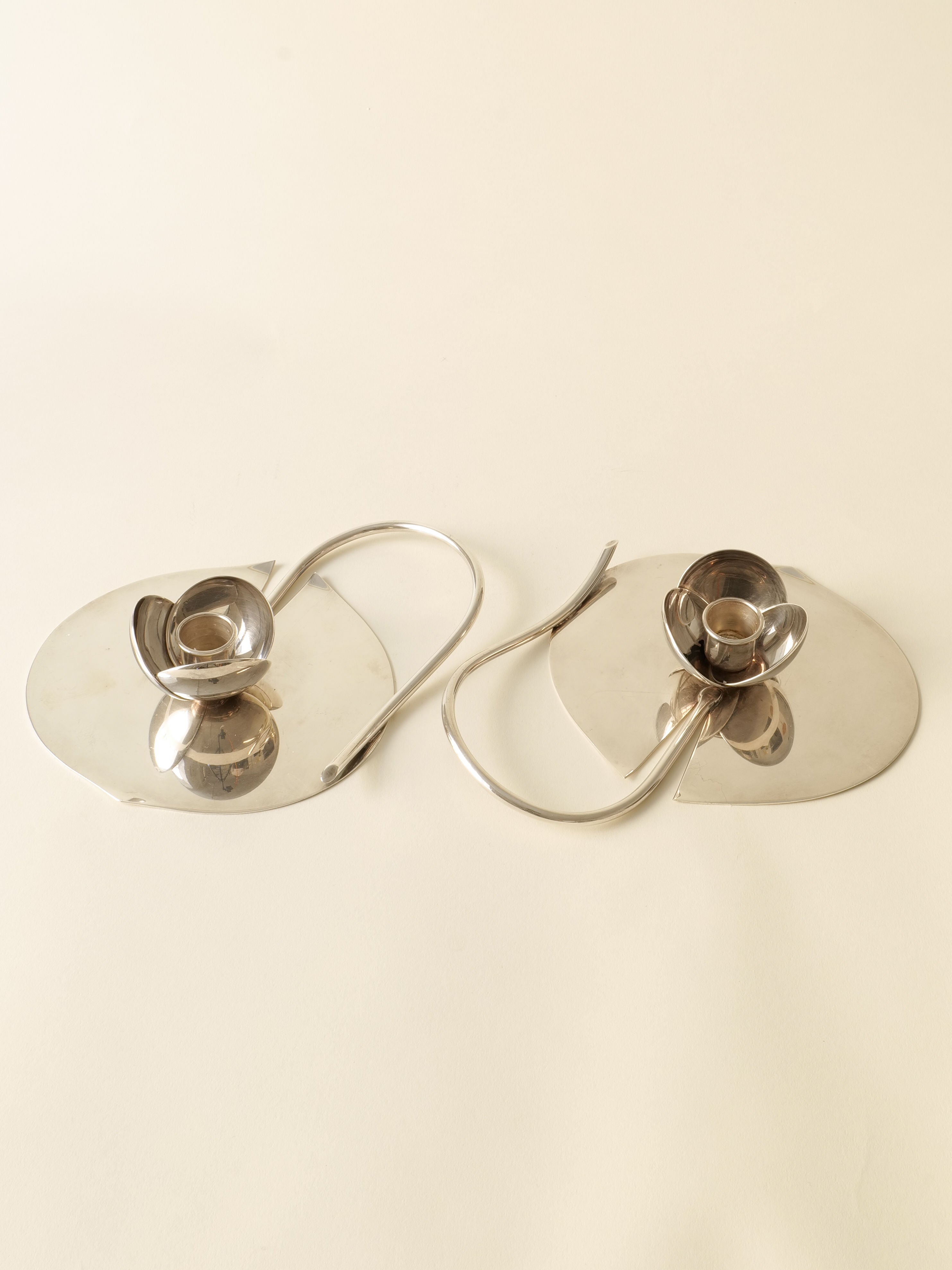 Two silver plated Candle Holder Lino Sabattini 1970s by Collection apart, each with a round base and curved handle, are placed side by side on a light surface. These Italian designer pieces feature abstract floral shapes.
