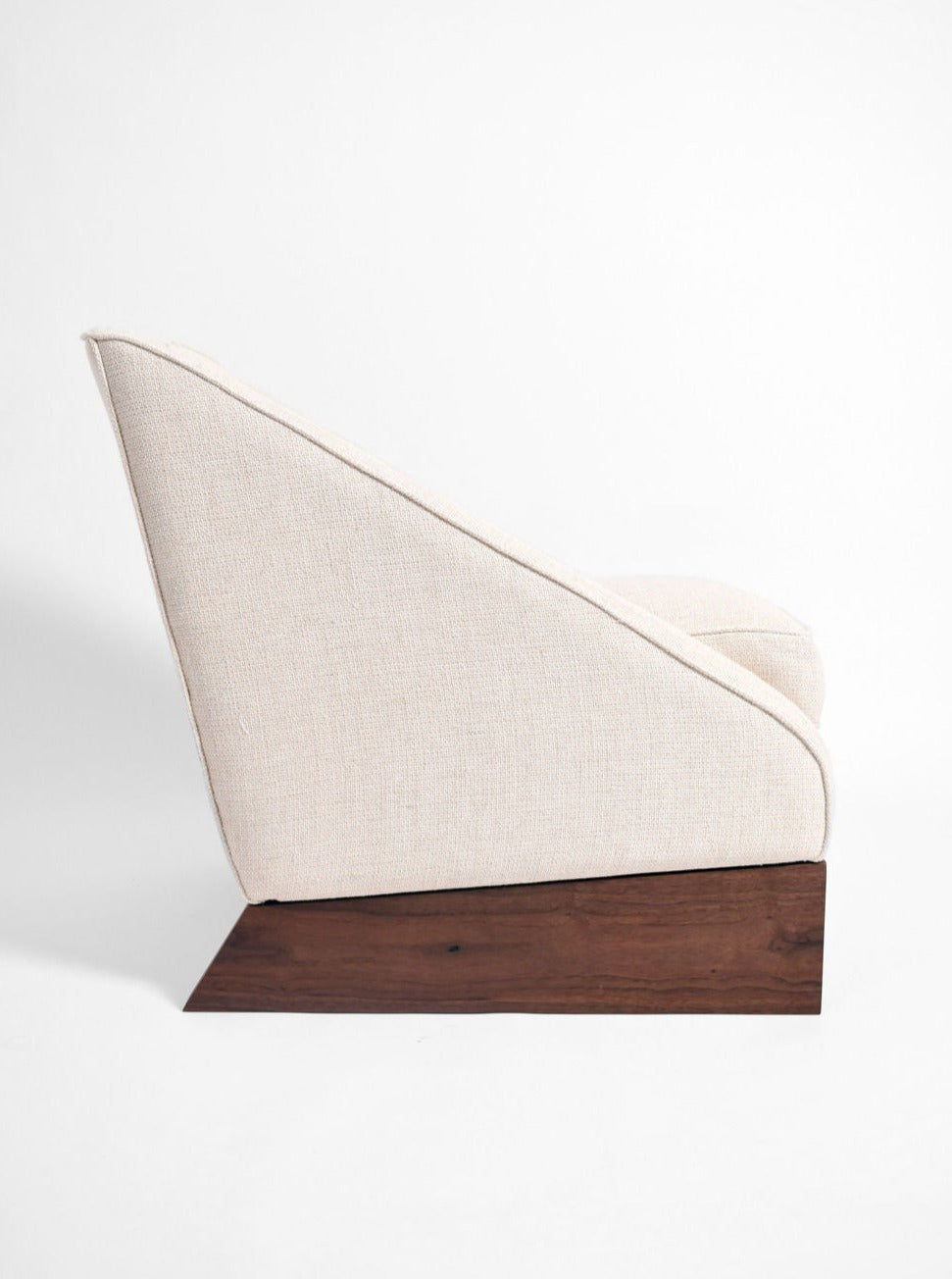 A side view of the Arrábida Armchair by Barracuda Interiors, showcasing its beige upholstery, slanted backrest, and wide flat wooden base, set against a plain white background.