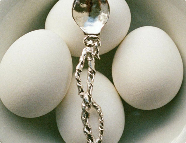 Close-up of four white eggs in a bowl with a silver twisted-handled spoon resting on top, creating a modern and artistic presentation.