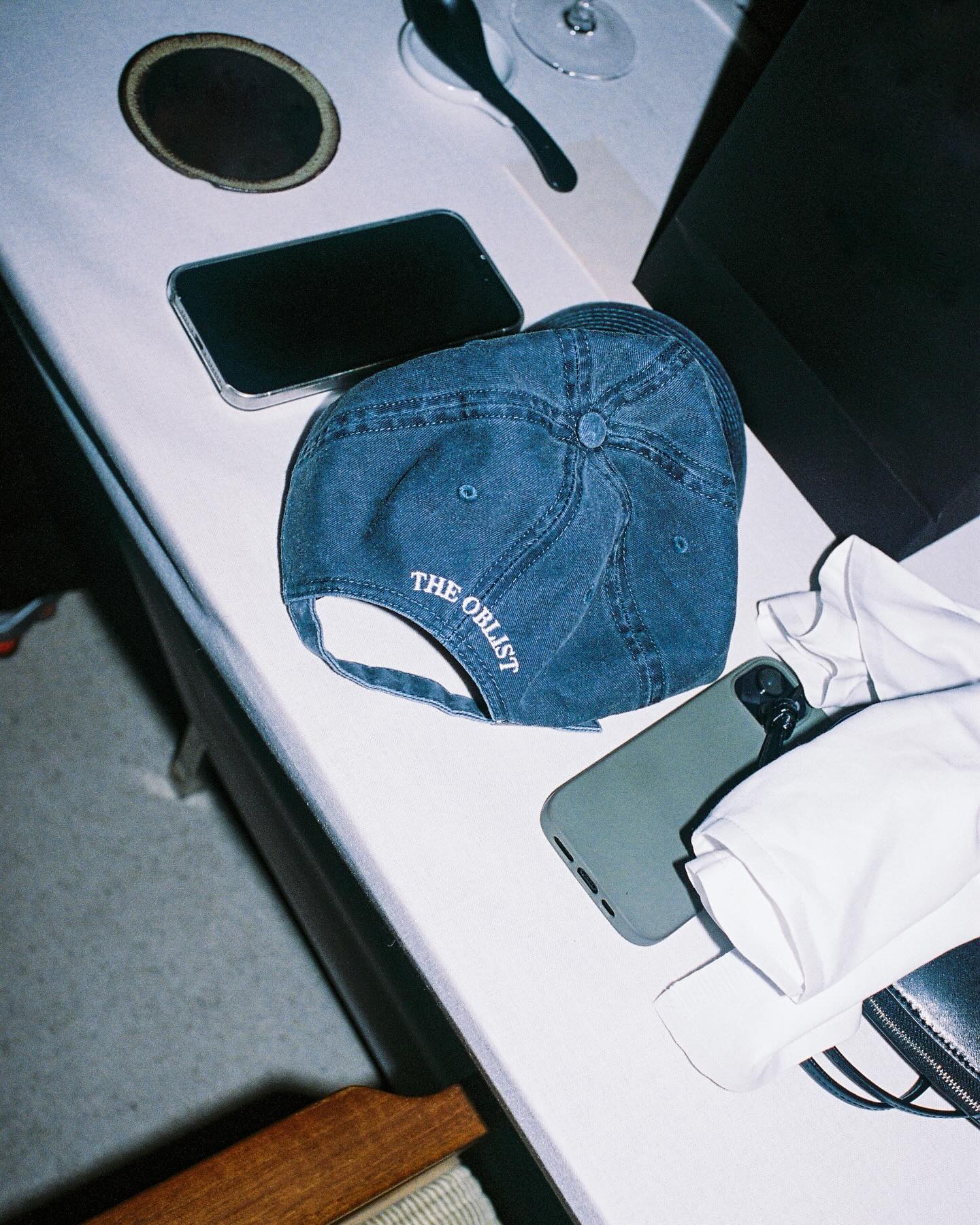 A smartphone rests beside The Singular Cap, a denim baseball cap by The Oblist, featuring "The Obelist" embroidered on it. It highlights its unisex fit and adjustable back strap. Accompanying the scene are a black phone case, white cloth, and coaster with a spoon, all contributing to an ambiance of distinctive taste in a casual and relaxed setting.