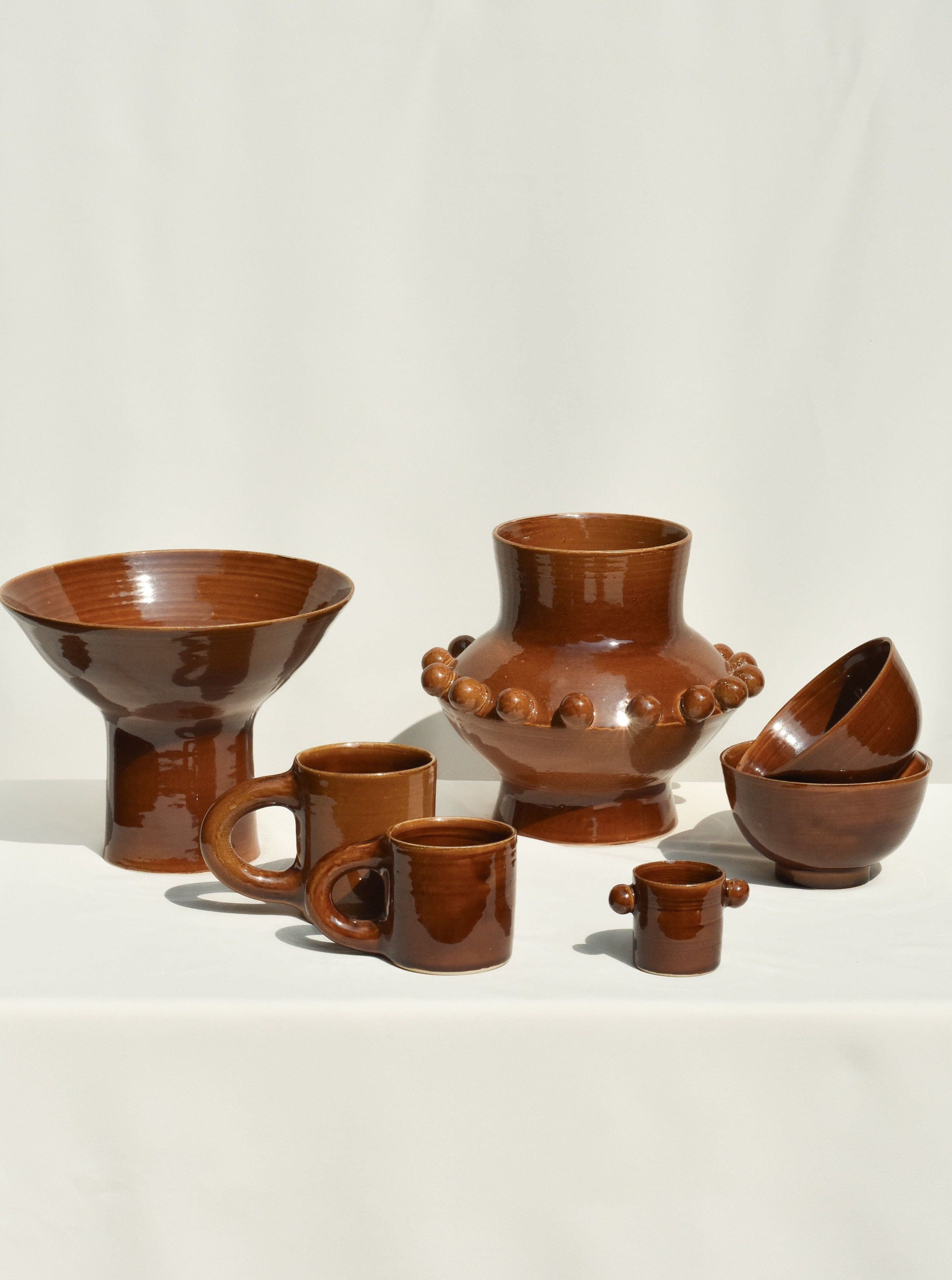 A selection of glossy brown ceramics from Landy Rakoto Ceramics, featuring cups, bowls, a wide-mouthed vessel, and the UFO Vase in Amber with round accents, all arranged on a light background.