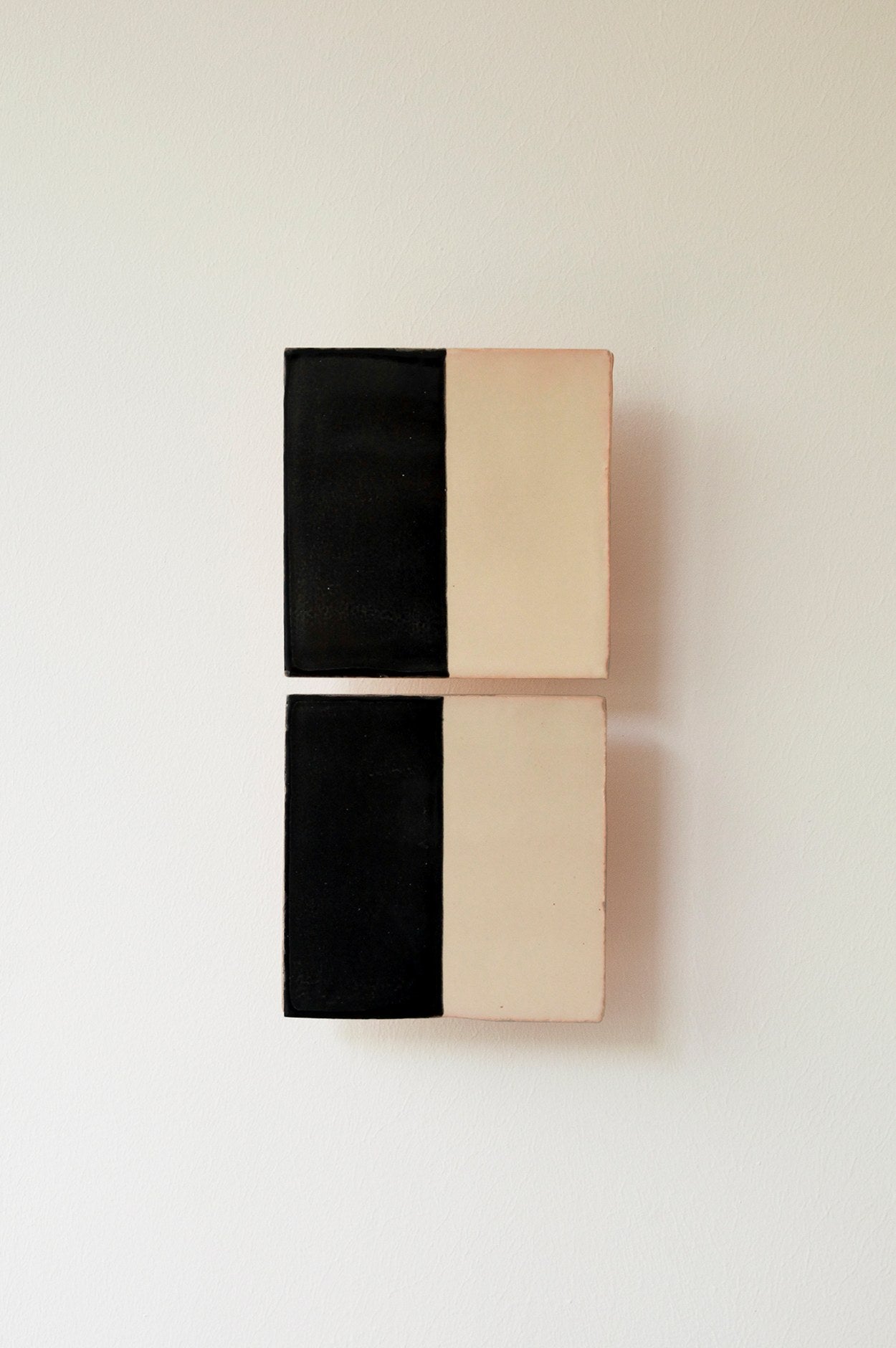 Two rectangular Tiles Line N by Violaine d'Harcourt, handmade and echoing ceramic wall art, are mounted on a white wall. Each piece is vertically split—black left, beige right—with divisions perfectly aligned.