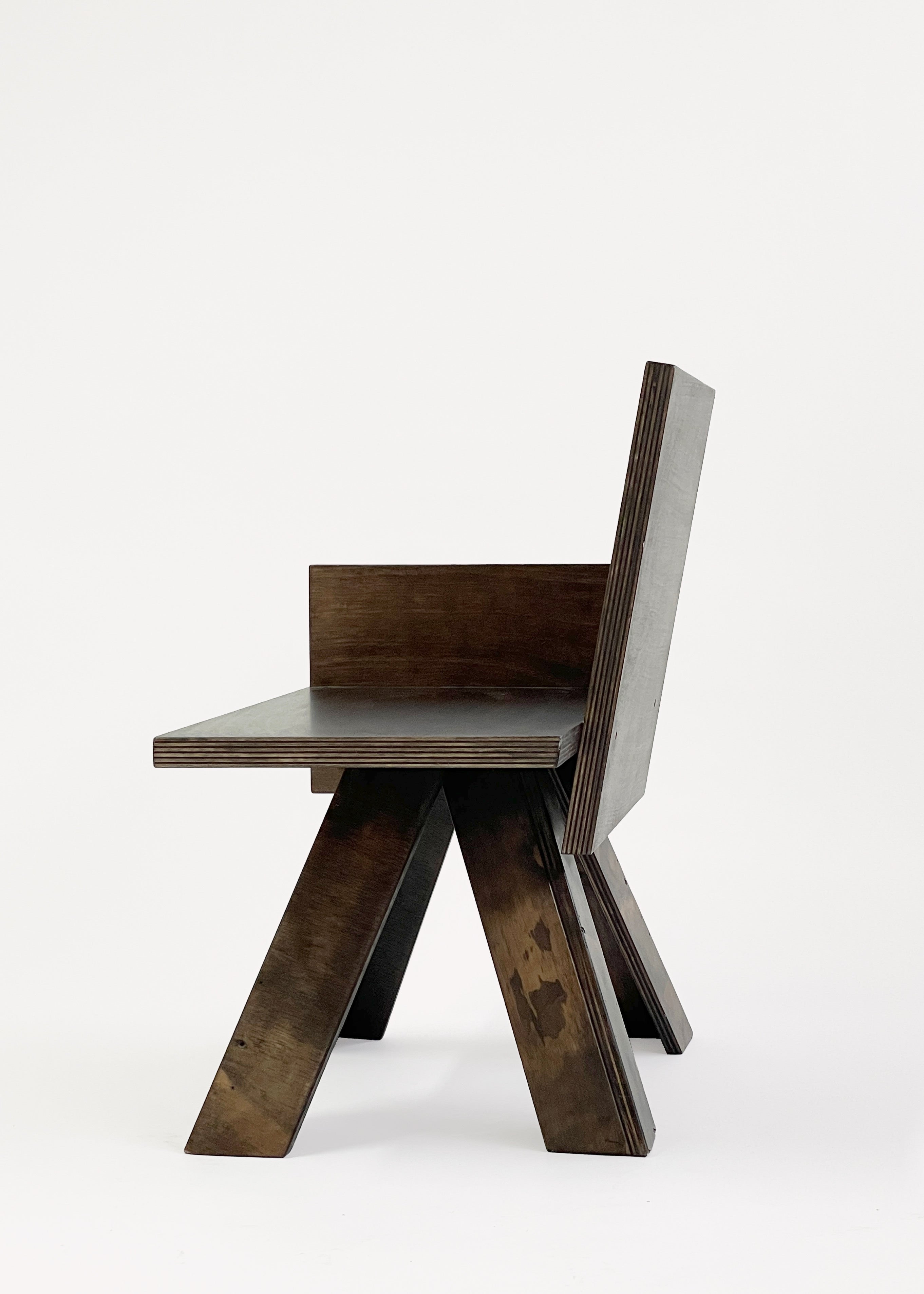 The End Table Chair by GOONS is a modern wooden chair with a geometric design, angled legs, flat seat, and straight backrest in dark wood—crafted by design studio Goons. This artisanal piece is shown on a plain white background.