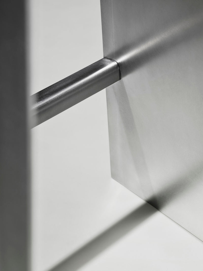 Close-up of the Camber Bar stool by Paul Coenen—minimalist metal design with clean lines and a smooth, brushed finish. Intersecting rectangular and cylindrical forms cast gentle shadows on a light background, reflecting modern furniture style.