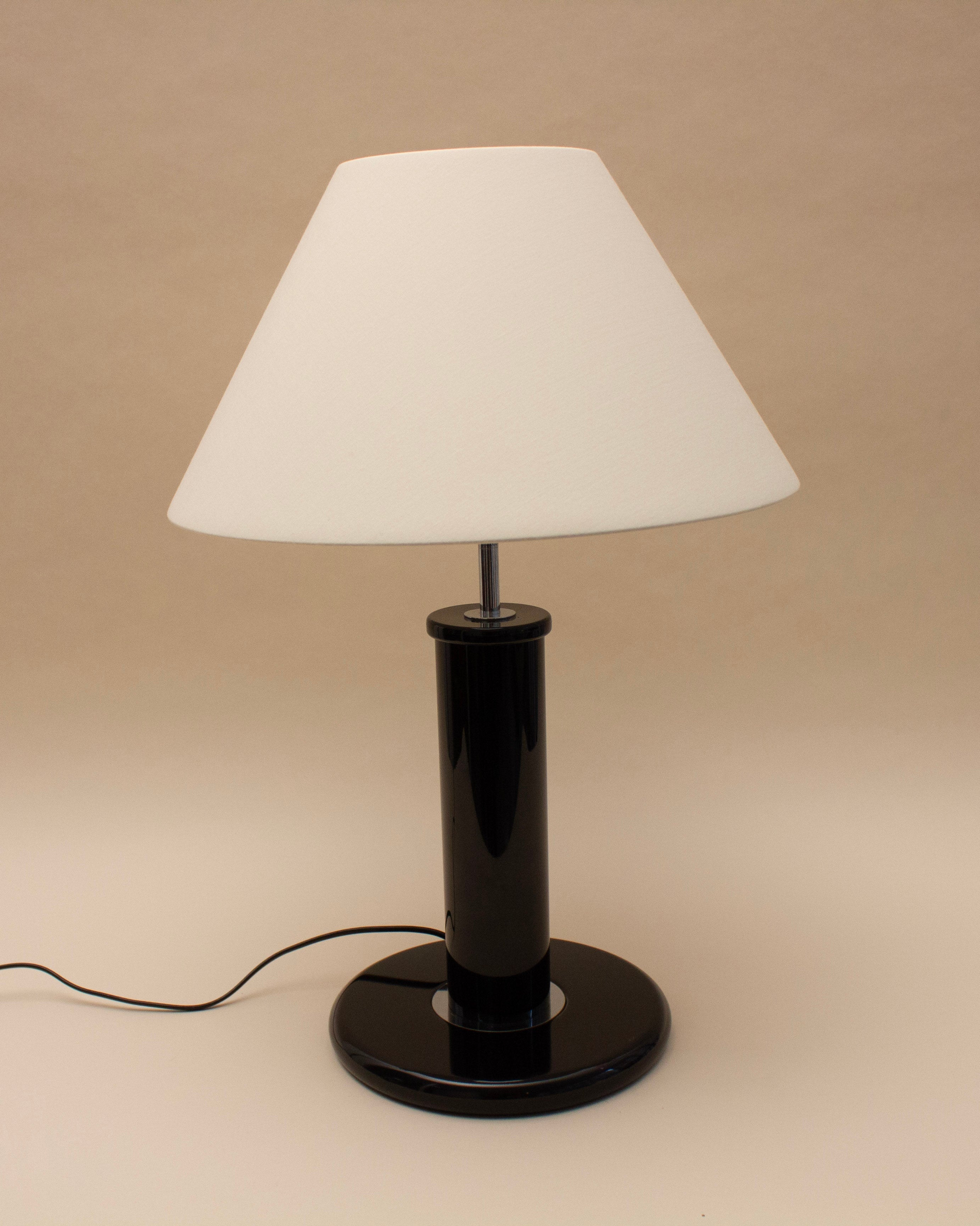 The SÒLER MADRID TABLE LAMP features a white conical shade and glossy black cylindrical base. It stands on a beige surface with its black power cord visible.