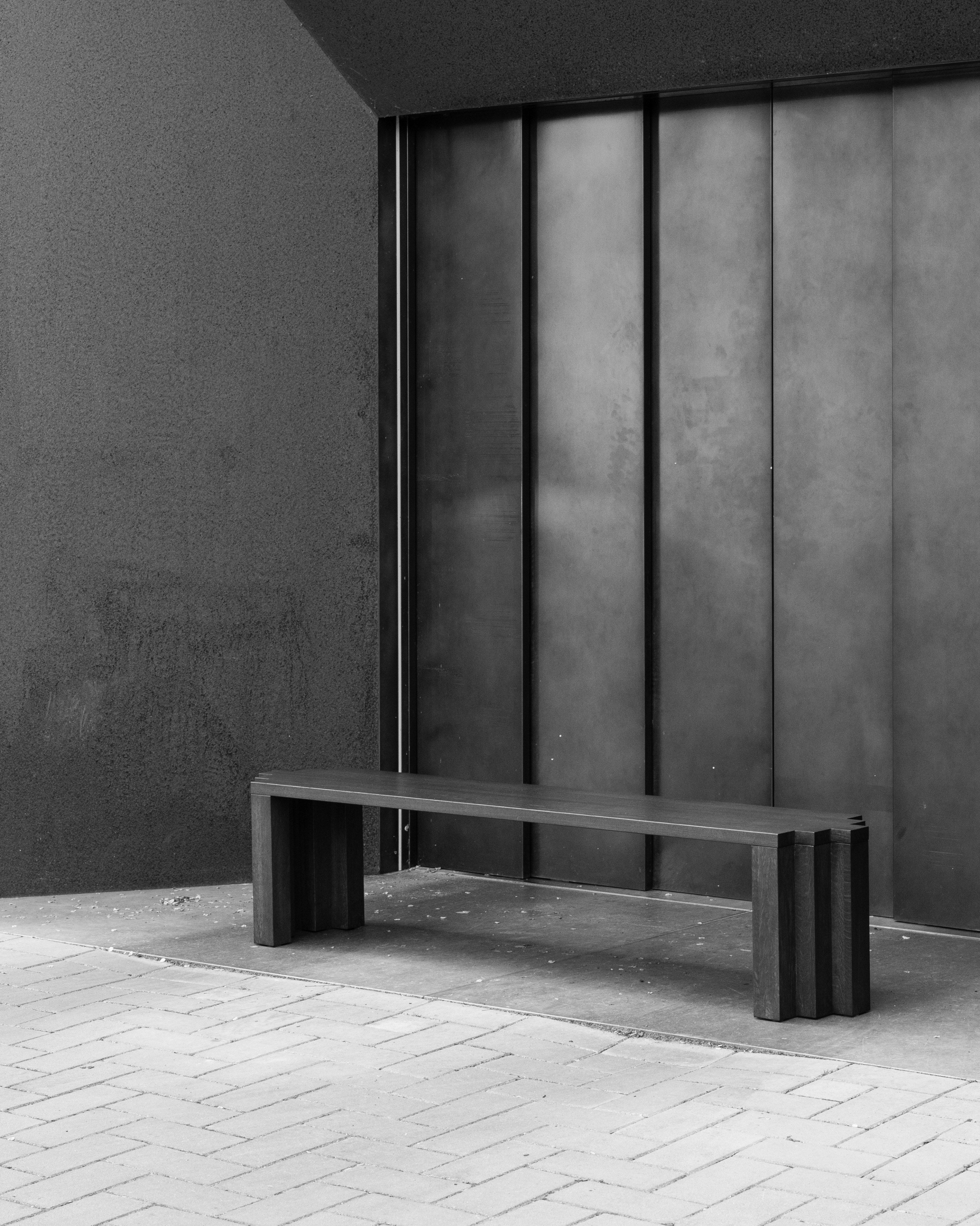 The Mokko Cadence Bench sits on a tiled pavement before a large, dark metal wall, creating a minimalist urban scene with monochromatic tones and solid European oak accents.