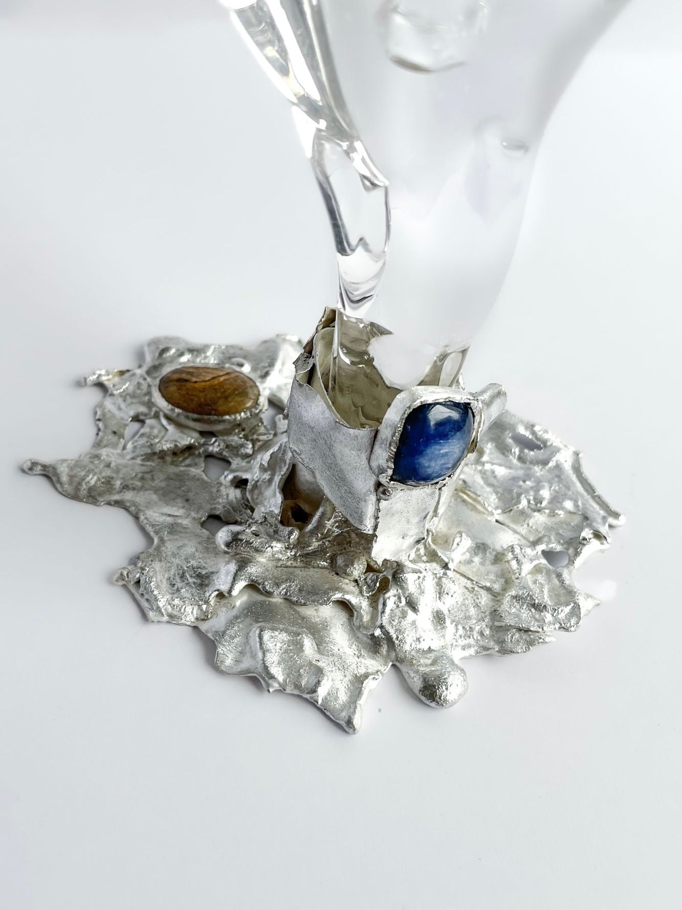 A close-up of the Jewelled Goblet no. 5 by Szkło Studio x Lindsey Fontijn highlights its sculptural silver base with melted edges and embedded blue and brown stones, supporting a hand-blown glass stem for striking contrast and texture.