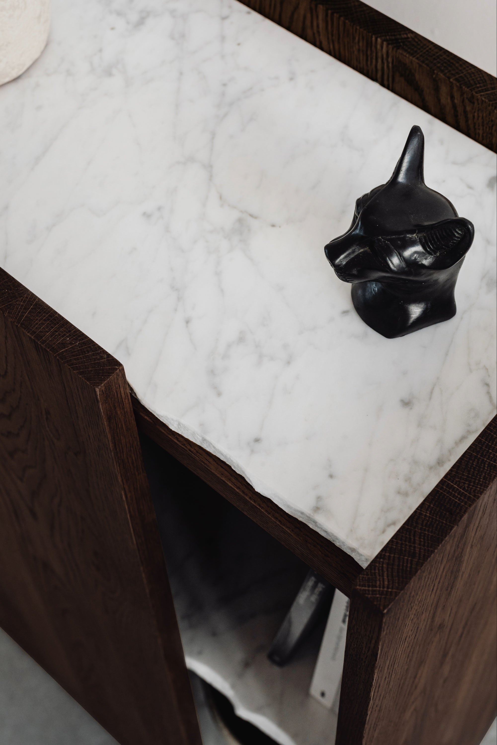 The PAVO Cabinet by un'common features a dark marble body with a white marble top. A small black animal head sculpture sits atop it, while the solid wood door is slightly open, showing papers stored inside.