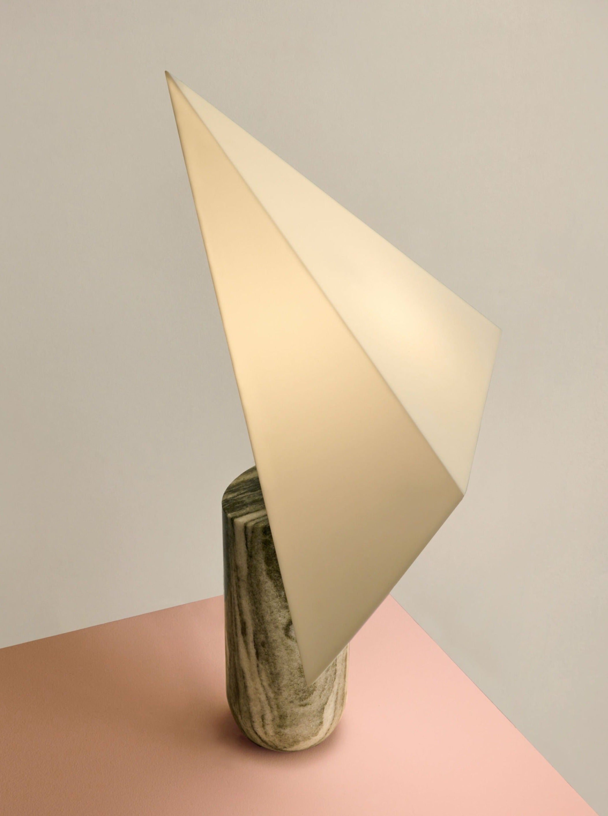 The Acme - Greek Marble table light by Mariza Galani features a handcrafted Greek marble cylindrical base and a large geometric white shade, displayed on a pink surface with a light beige background.