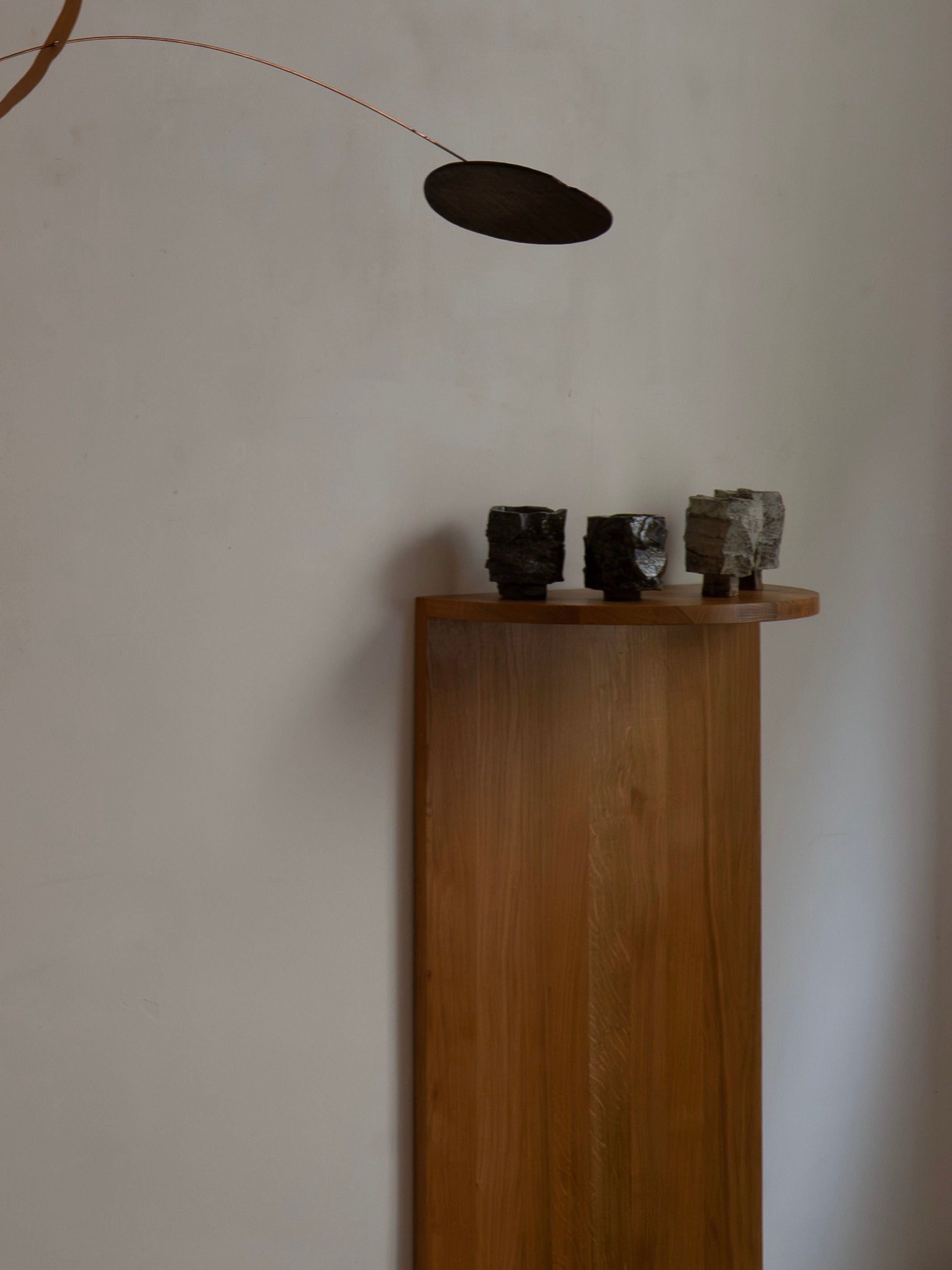 A handcrafted solid oak Megaron Minimalist Shelf by COSEINCORSO displays three textured ceramic cups against a plain wall. Above, a thin metal rod suspends a flat, oval black mobile for an artistic accent.