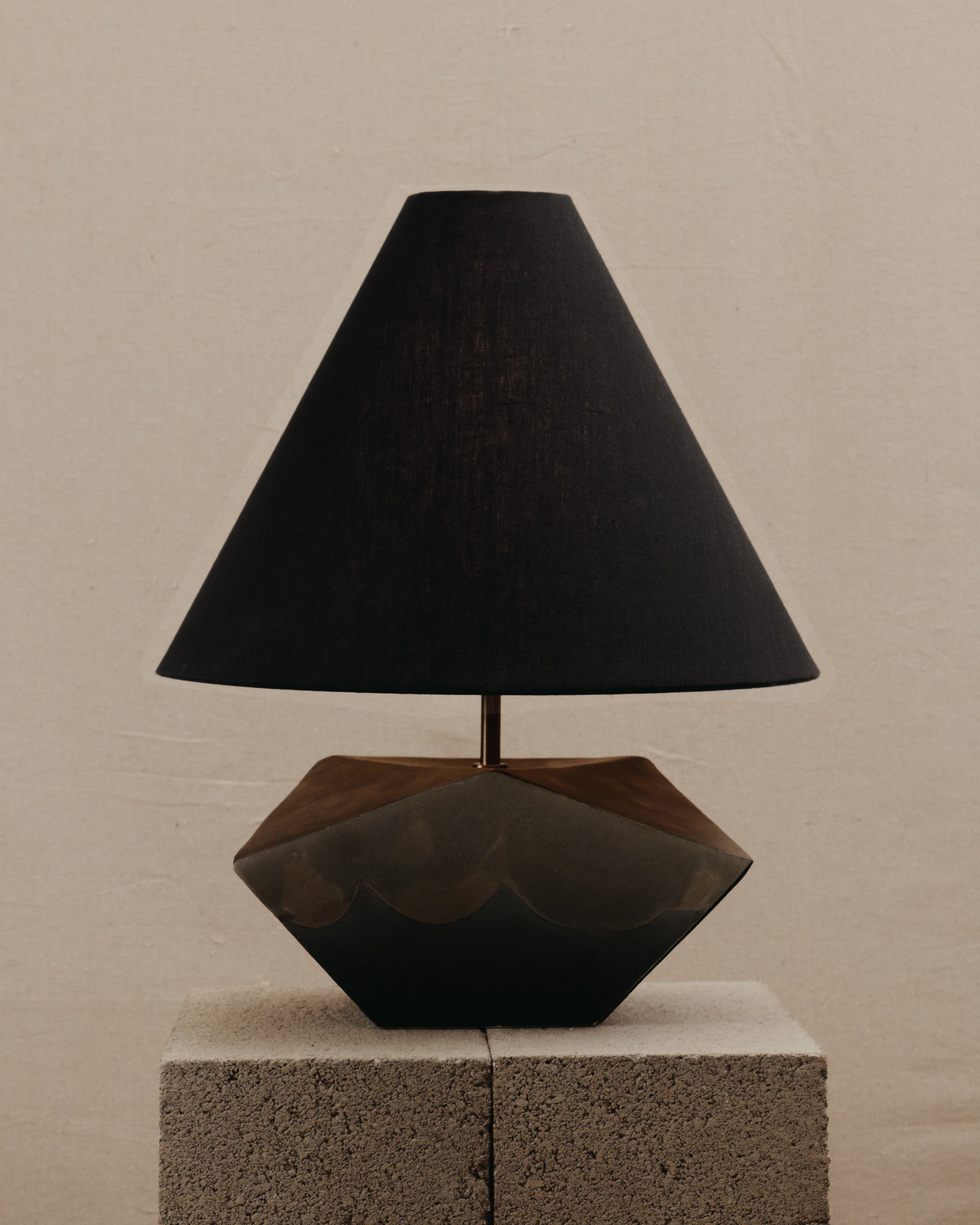 The Summit Table Lamp by JS/WRKSHP features a geometric dark base and wide linen shade, displayed on stacked concrete blocks to highlight its organic modernist style against a plain beige background.