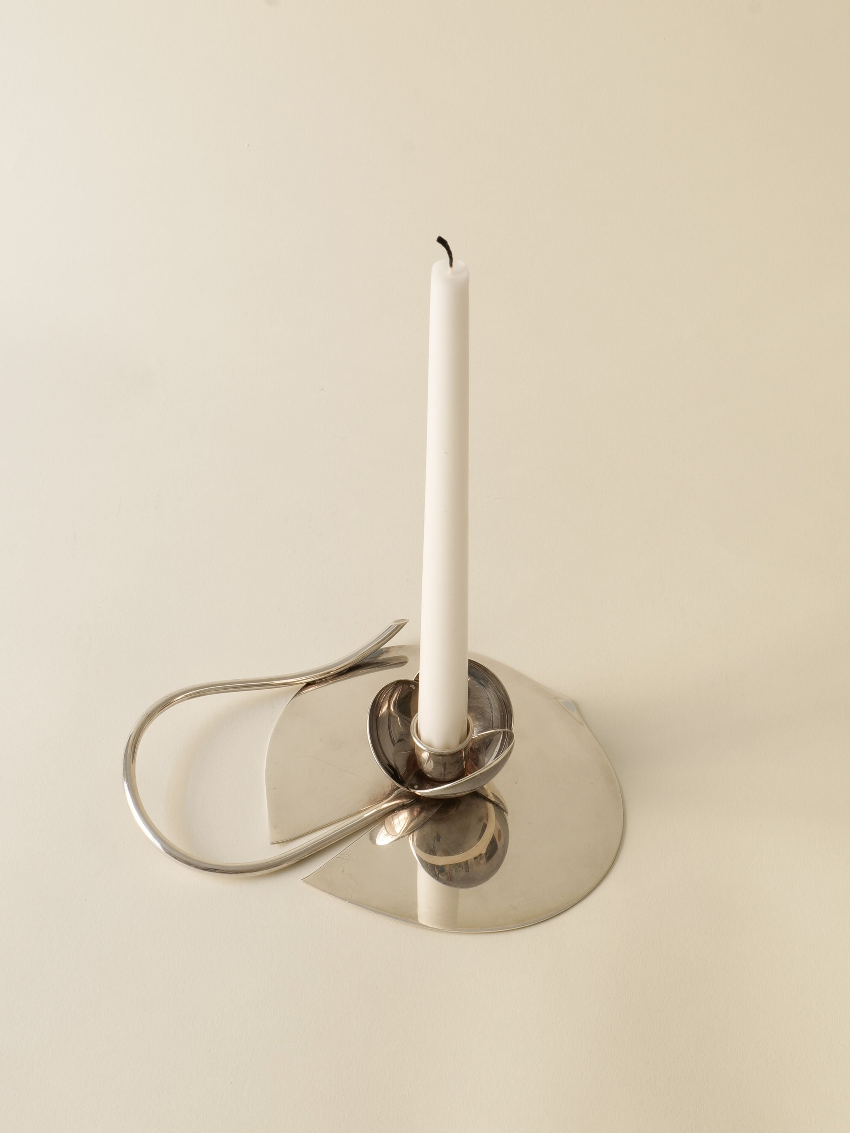 A white candle sits in the minimalist, reflective Candle Holder Lino Sabattini 1970s by Collection apart, featuring a silver-plated curved handle on a light surface.