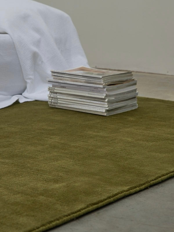 Hand-woven Rug in New Zealand Wool, Olive Green — side view with magazines stacked nearby