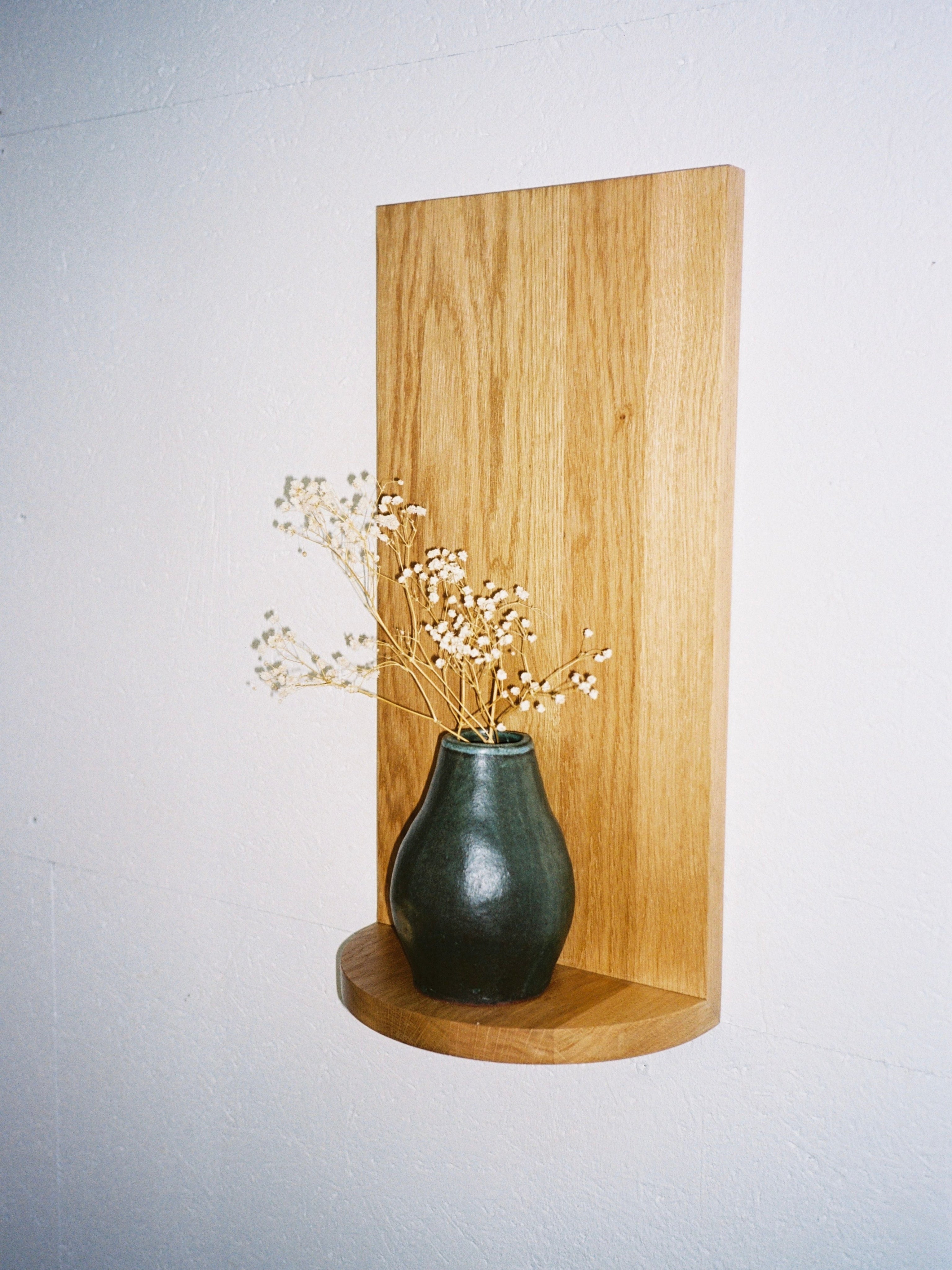 A small green ceramic vase with dried white flowers rests on The Megaron - Minimalist Shelf in Wood (Small) by COSEINCORSO, handcrafted and mounted on a white wall.