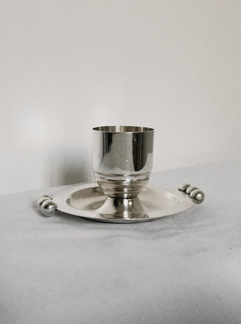 The Manushi Silver Plated Detail Egg Cup rests on a matching round tray with decorative handles, displayed on a soft, light surface against a plain background.