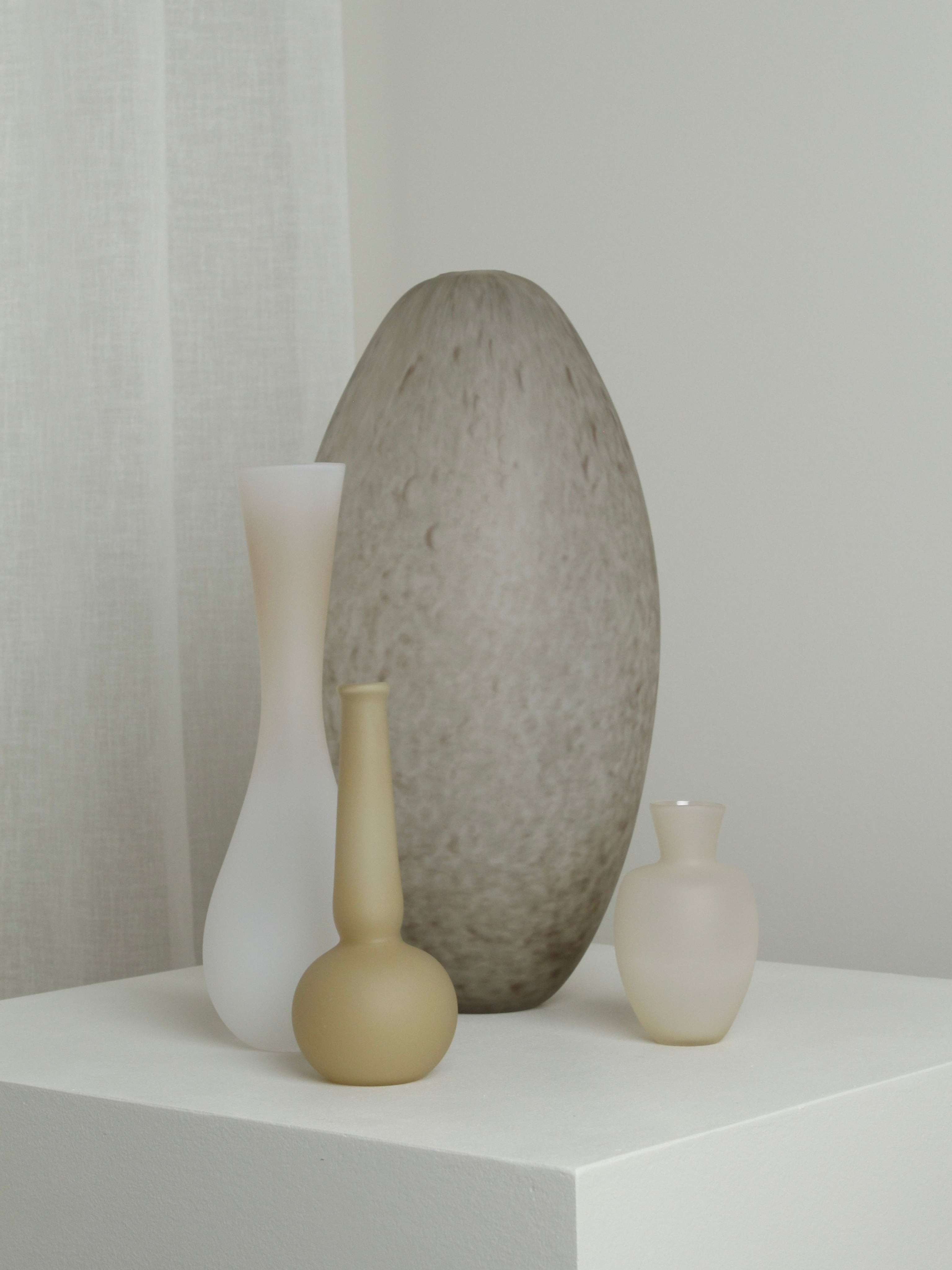 The Small Vase in Caramel by Verre d’Onge sits on a white pedestal among neutral minimalist vases, its warm tone and smooth, refined shape standing out against the light gray wall and white curtain.