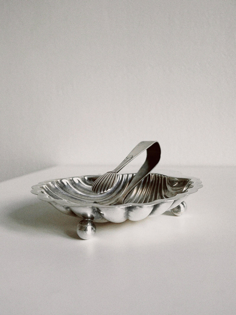 The Manushi Shell Shaped Dish and Prong features a silver shell-inspired dish with round feet, holding matching prong tongs. The set is displayed on a white surface with a clean, light background.